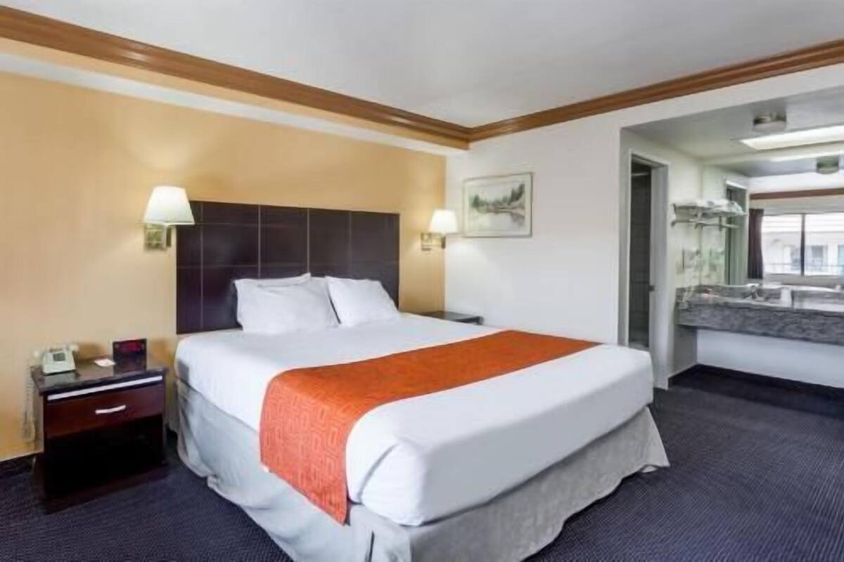 A hotel room with a bed, bedside tables, a nightstand, a lamp, a painting, and a bathroom visible through an open door.
