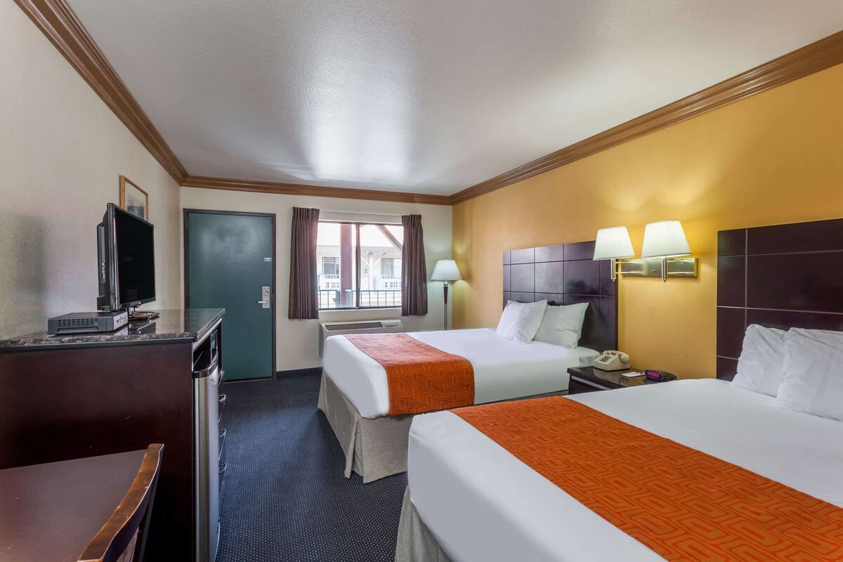 A hotel room with two beds, a desk with a television, a bedside table with a lamp, and a window with curtains.