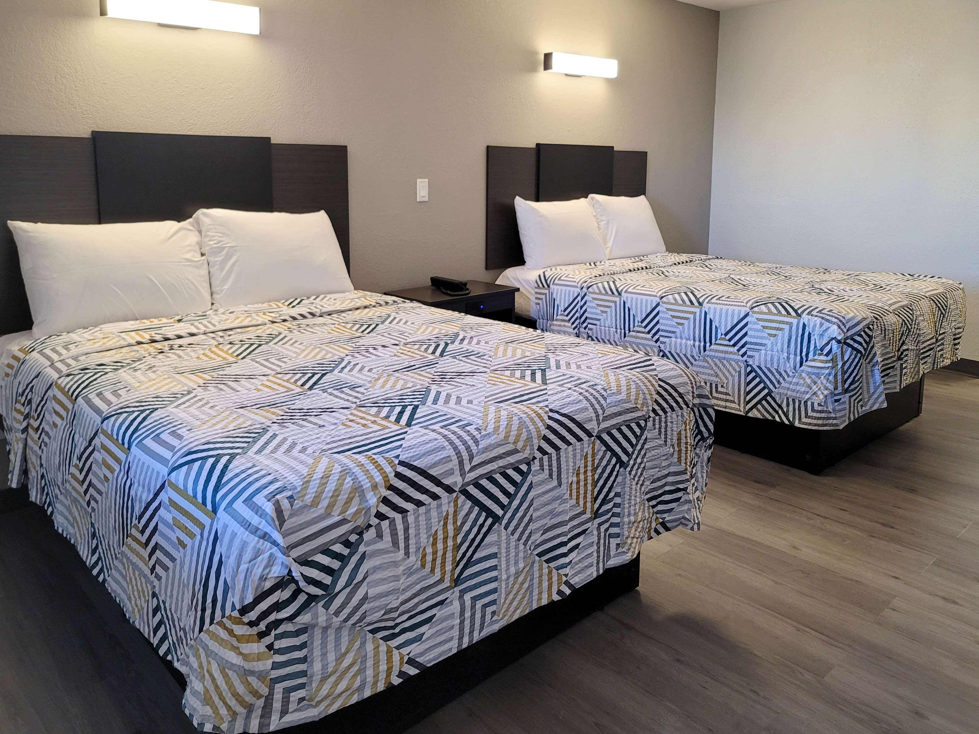Hotellihuoneessa on kaksi sänkyä kuviollisilla vuodevaatteilla, yöpöytä ja seinään kiinnitetyt valaisimet.