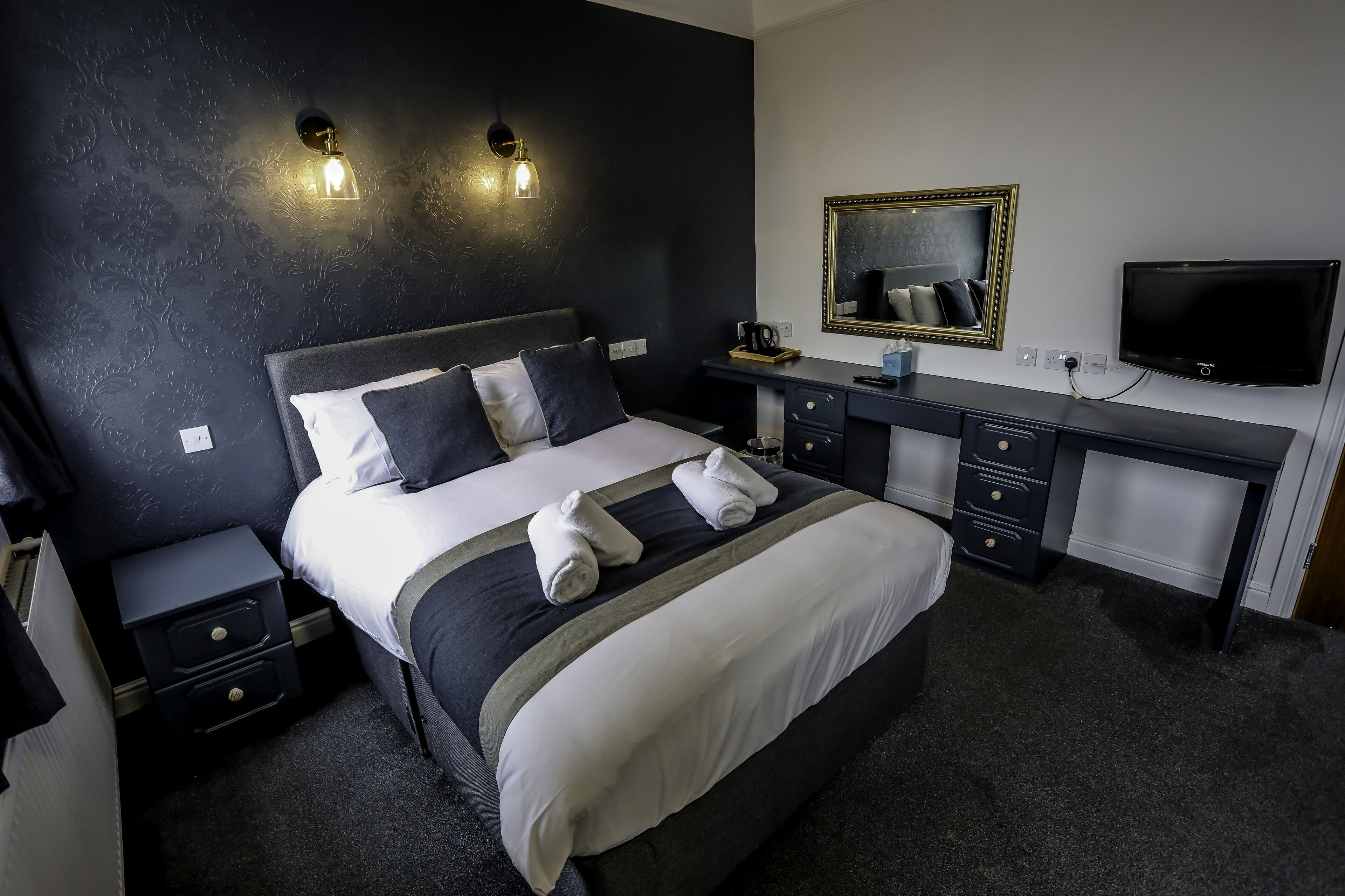 A hotel room with a large bed, bedside tables, a desk, a mirror, and a TV.