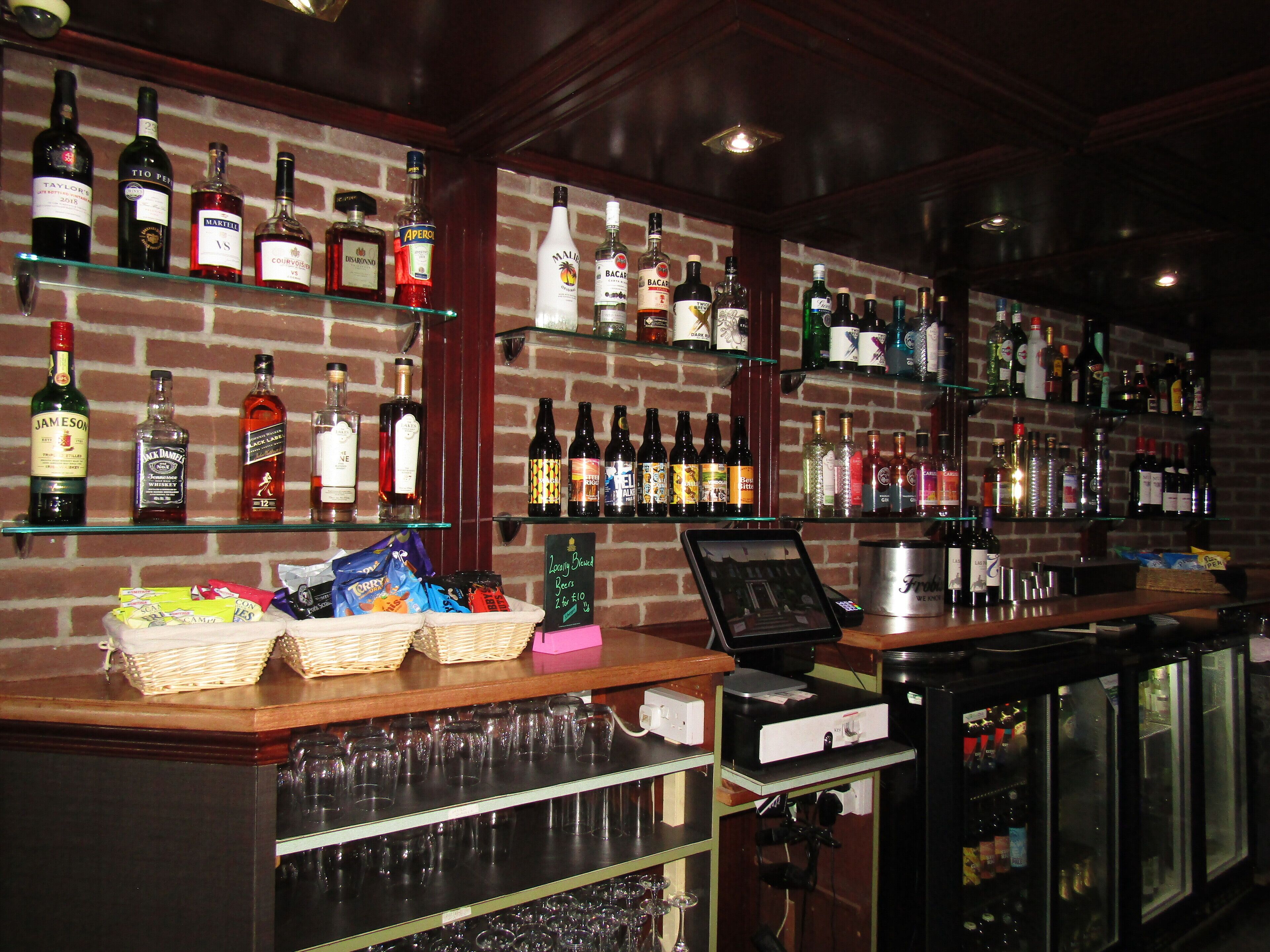 A bar area with shelves of various liquor bottles, a cash register, and a selection of snacks and glasses.
