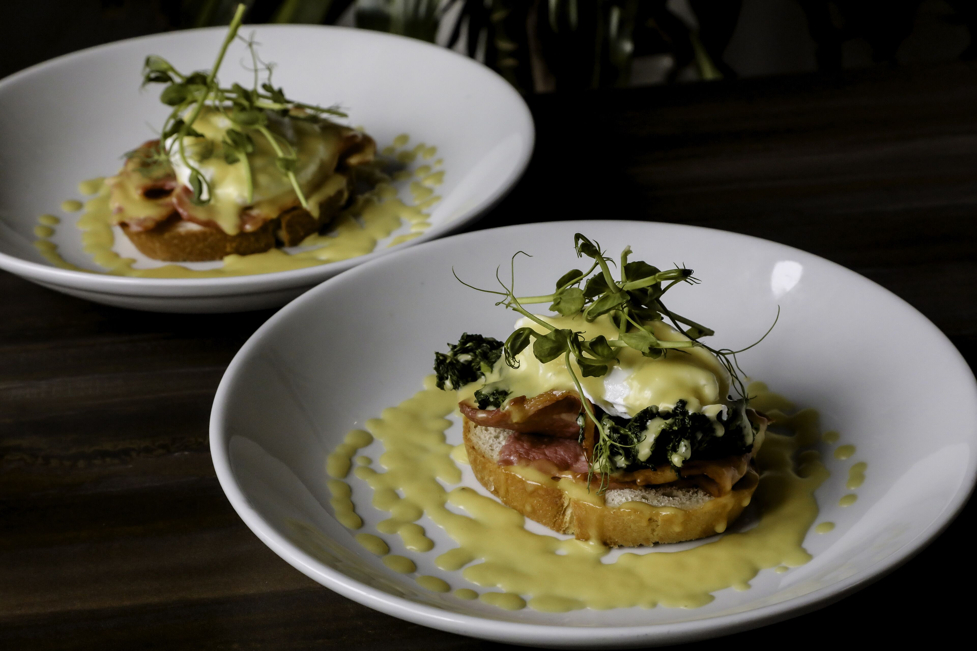 Two white plates with a dish consisting of greens, egg, and meat on a slice of bread, accompanied by a yellow sauce.
