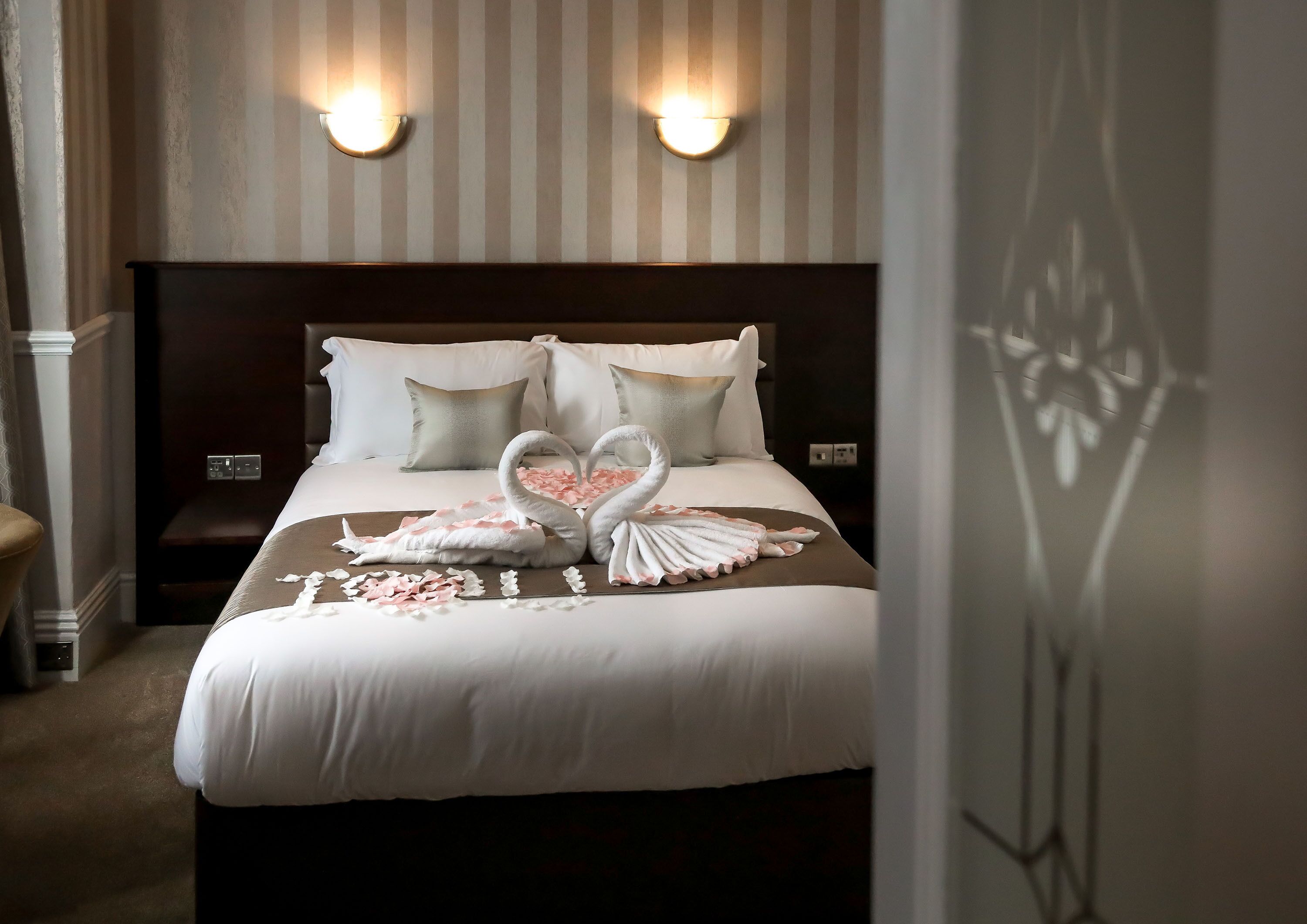 Una cama impecablemente hecha, adornada con una toalla en forma de corazón y pétalos de rosa, un cabecero de madera oscura y dos luces fijadas a la pared.