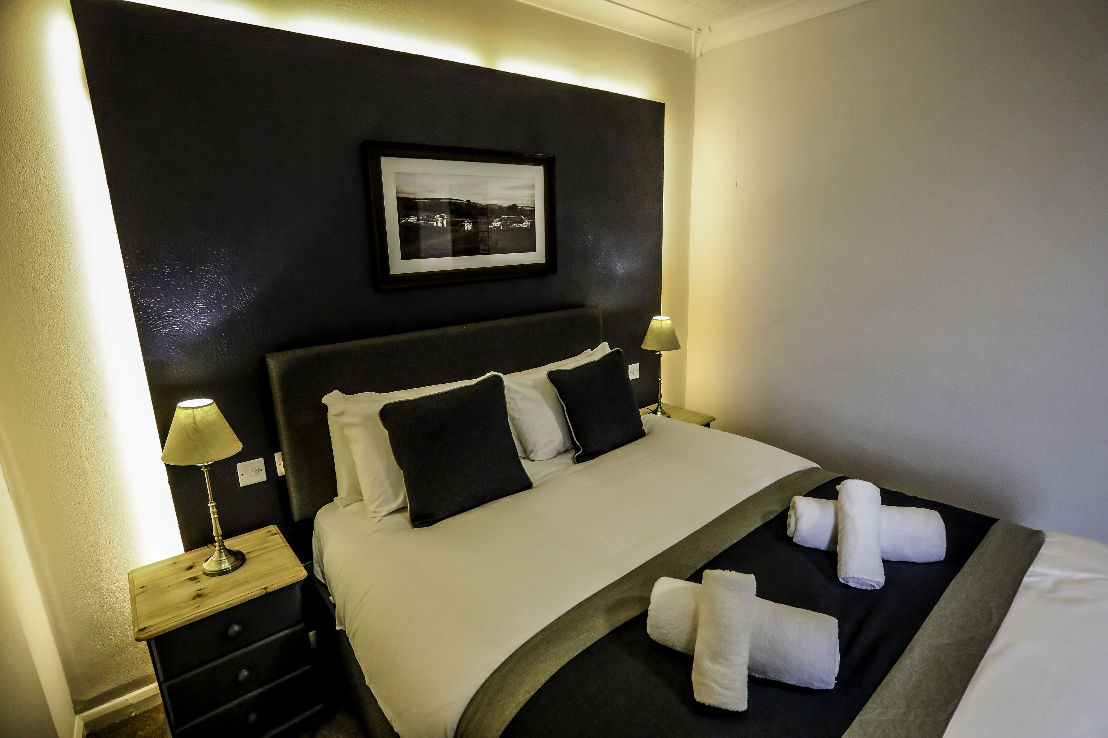 Habitación de hotel con una cama bien hecha, dos lámparas de noche, un cuadro en la pared y una mesita con lámpara.