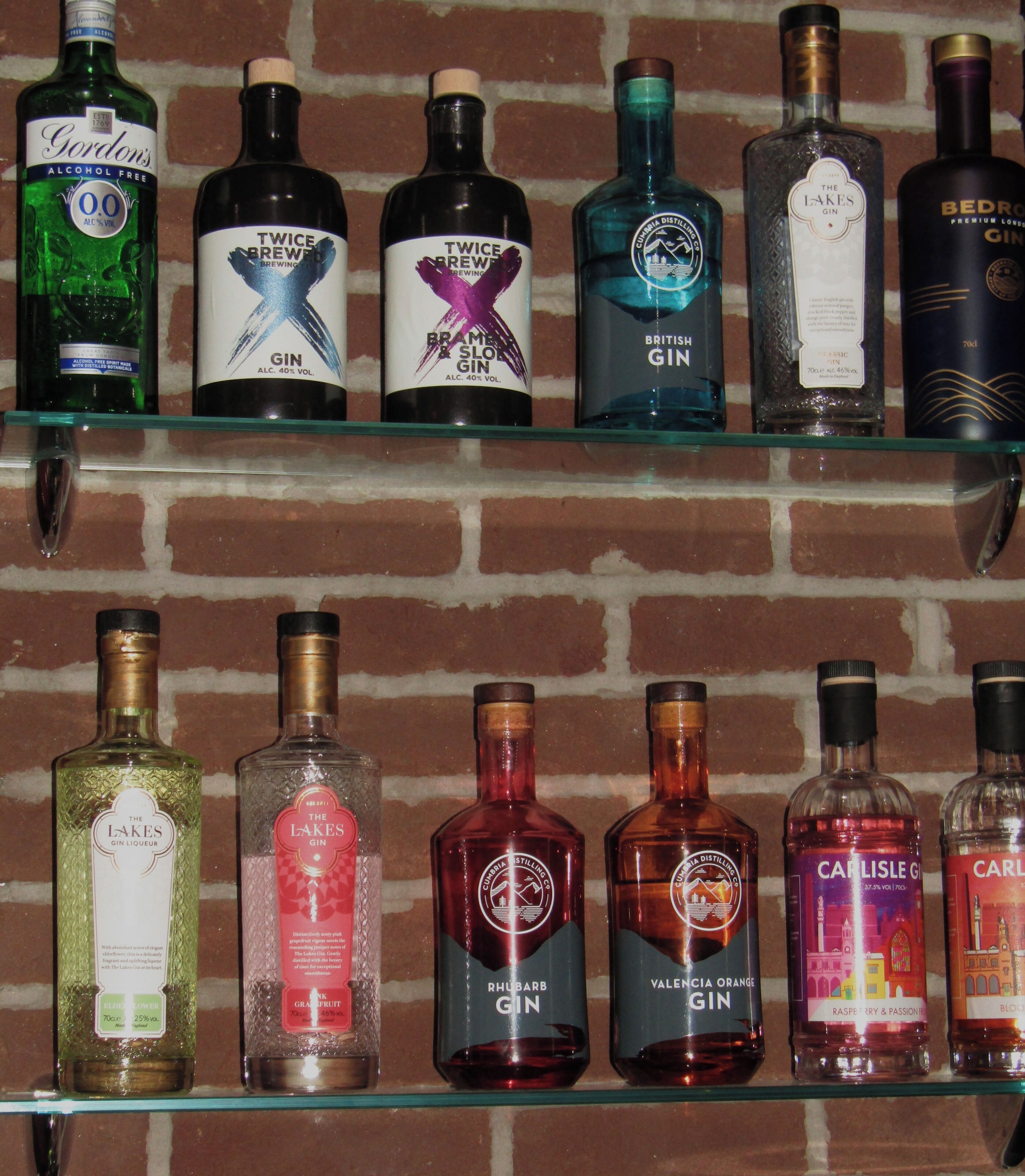 Diversas garrafas de gin posicionadas em uma prateleira de vidro, com uma parede de tijolos ao fundo.