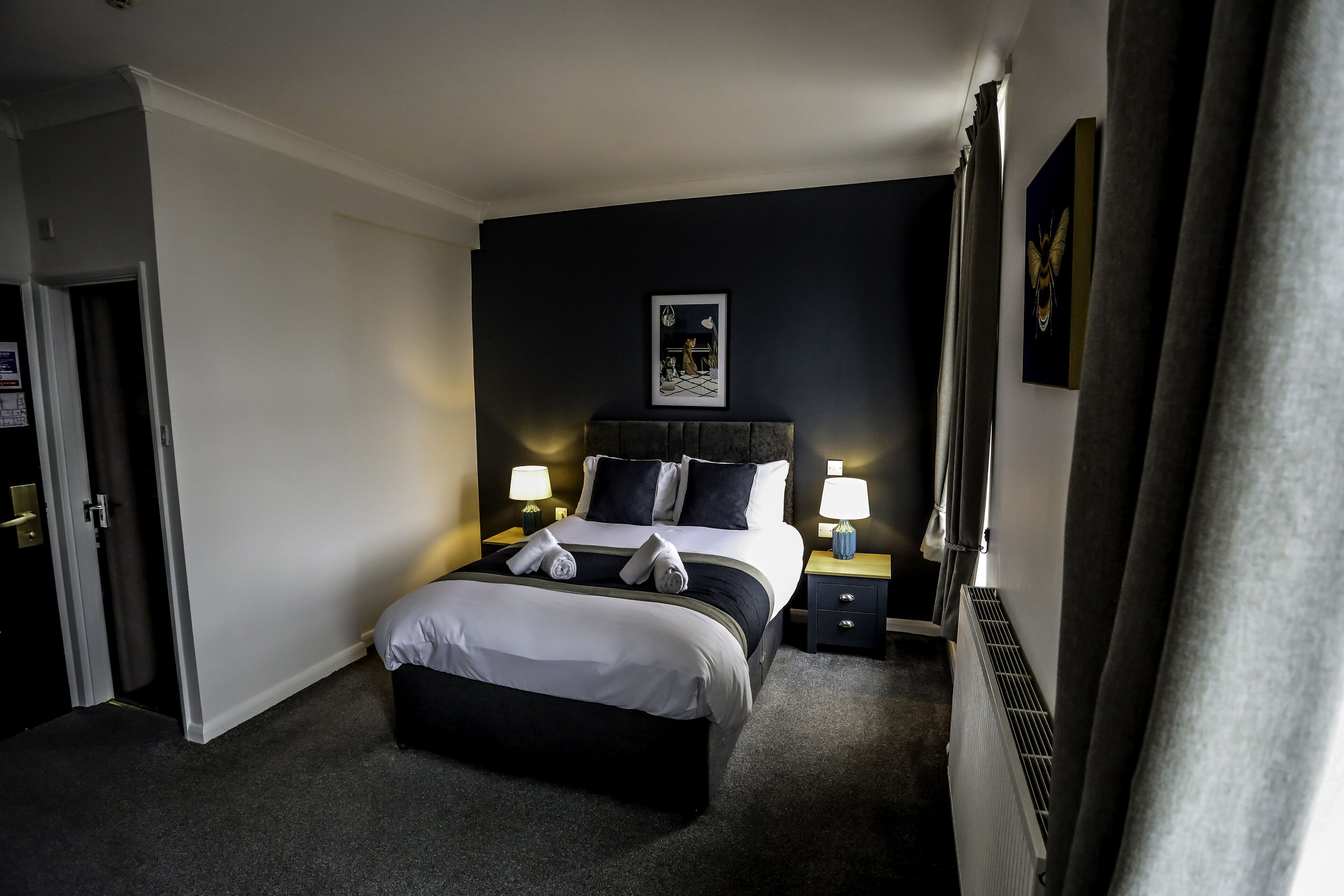 Ein Hotelzimmer mit einem großen Bett, zwei Nachttischen mit Lampen, einer Kopfteil und einem gerahmten Bild an der Wand, sowie einem Heizkörper.