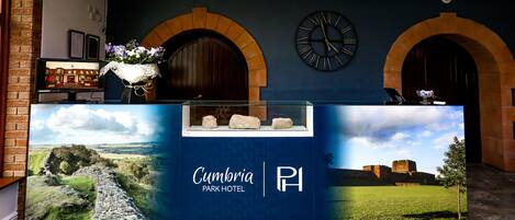 Una recepción en el Cumbria Park Hotel, con un gran cartel del nombre y logotipo del hotel, dos relojes sobre el mostrador y un arreglo floral a la izquierda.