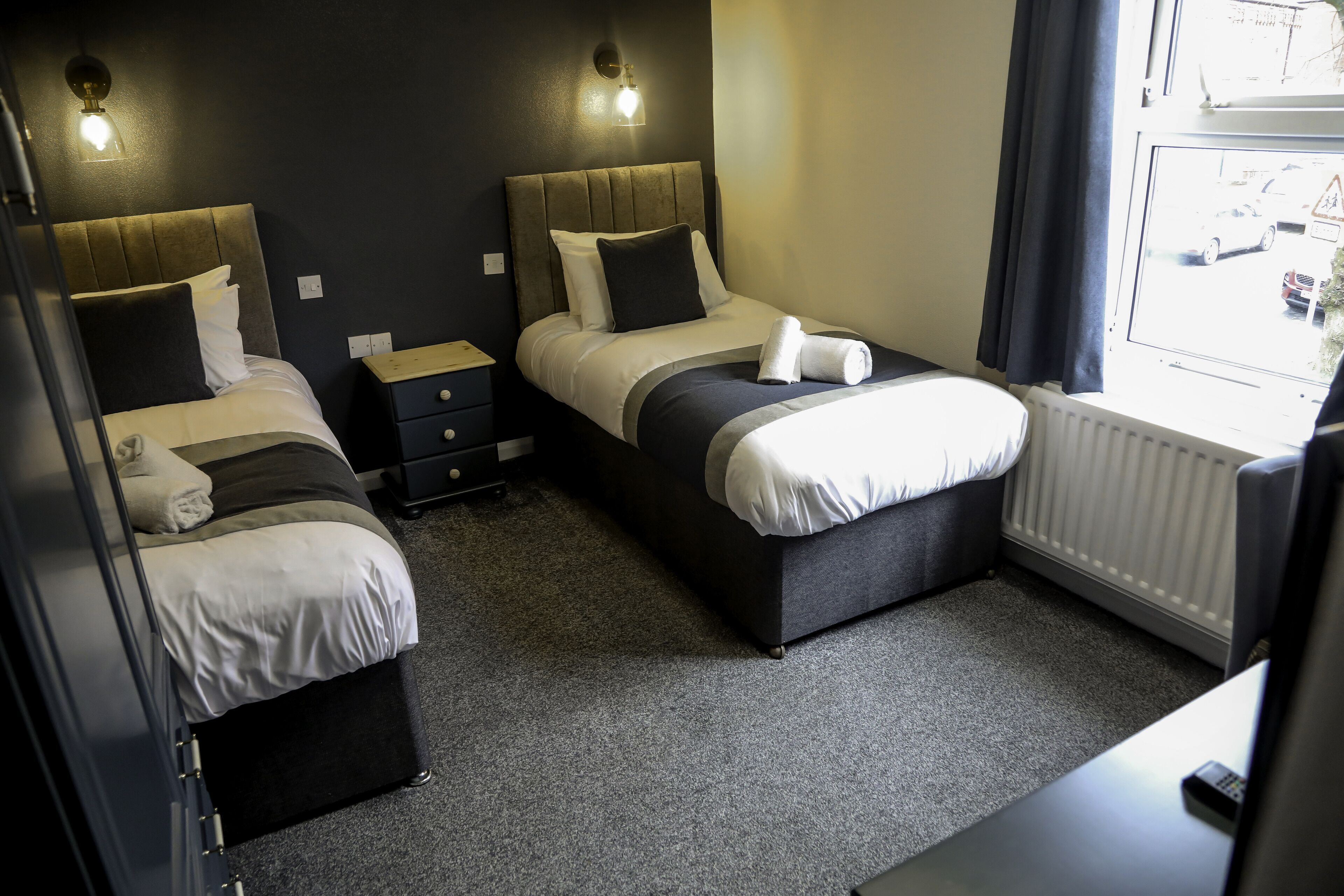 A hotel room with two beds, a small bedside table, a nightstand, and a window with a view of the street.