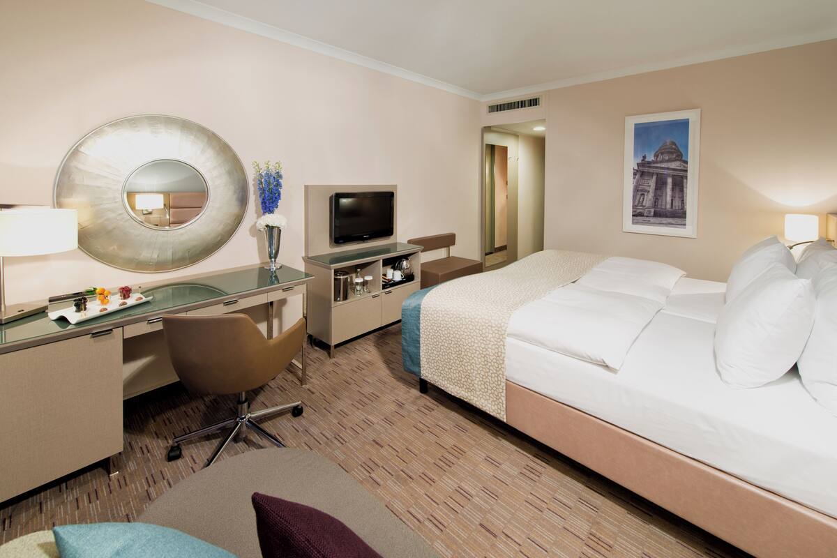 Habitación de hotel con cama, escritorio con televisor, silla, espejo y un cuadro en la pared.