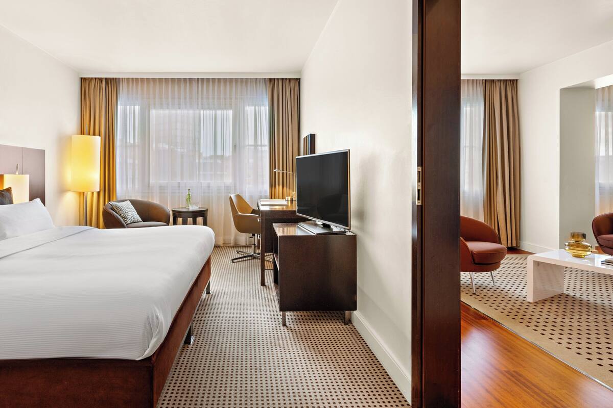 Habitación de hotel con una cama grande, un escritorio con televisor, una silla y una mesita de noche con lámpara.