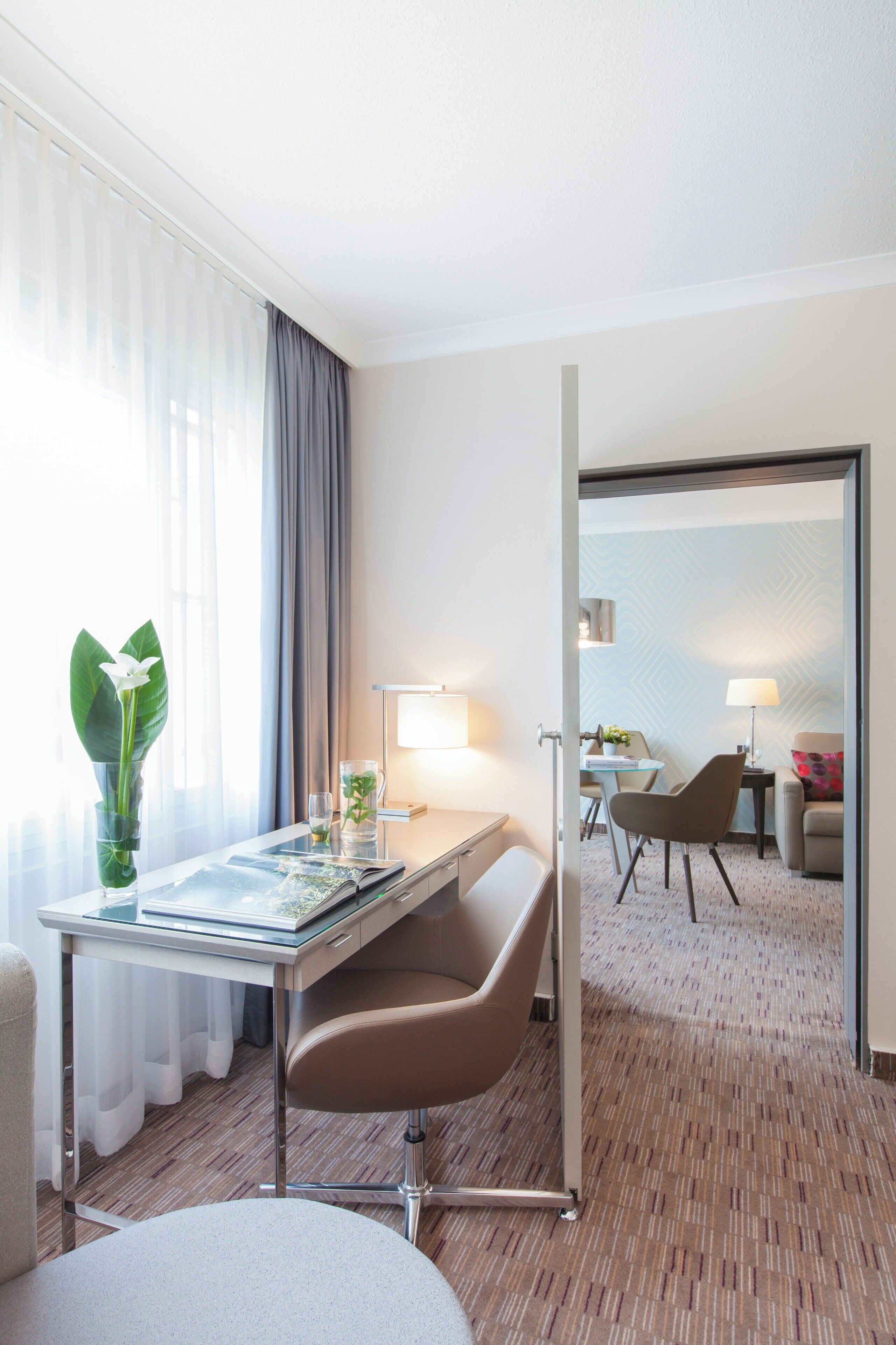 A hotel room with a desk, chair, lamp, and a view of the adjacent room.