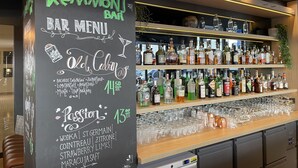 Un menu de bar proposant une variété de boissons et de prix, un comptoir de bar avec des étagères remplies de bouteilles d’alcool, et un espace bar équipé de tabourets.