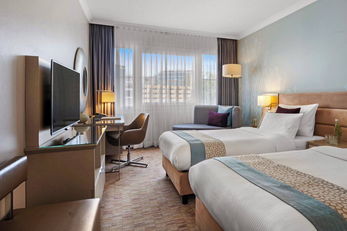 Habitación de hotel con dos camas, un escritorio con televisor, una silla y vistas a la ciudad a través de amplias ventanas.