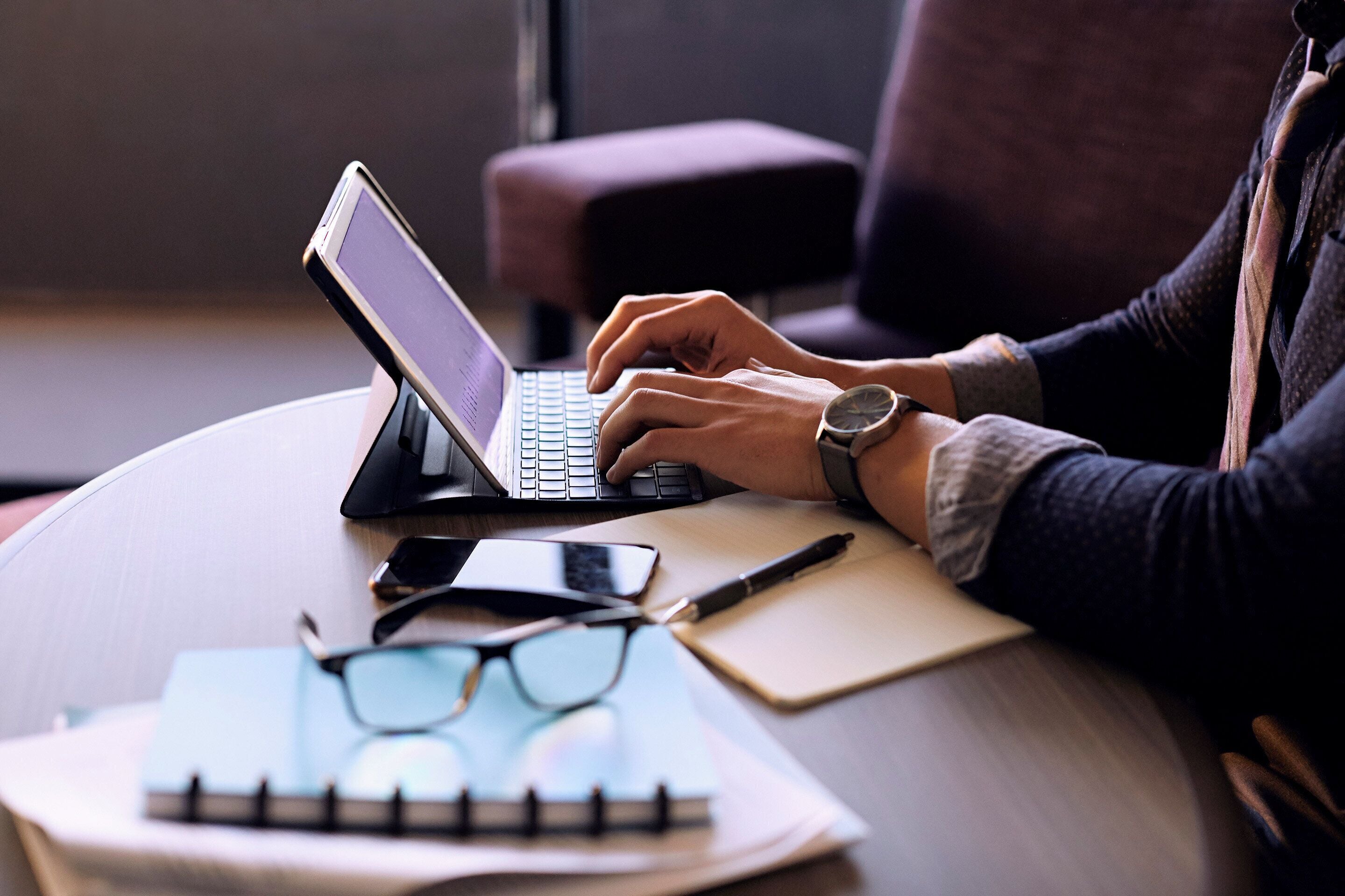 A person using a laptop on a round table with a notebook, glasses, and a pen.
