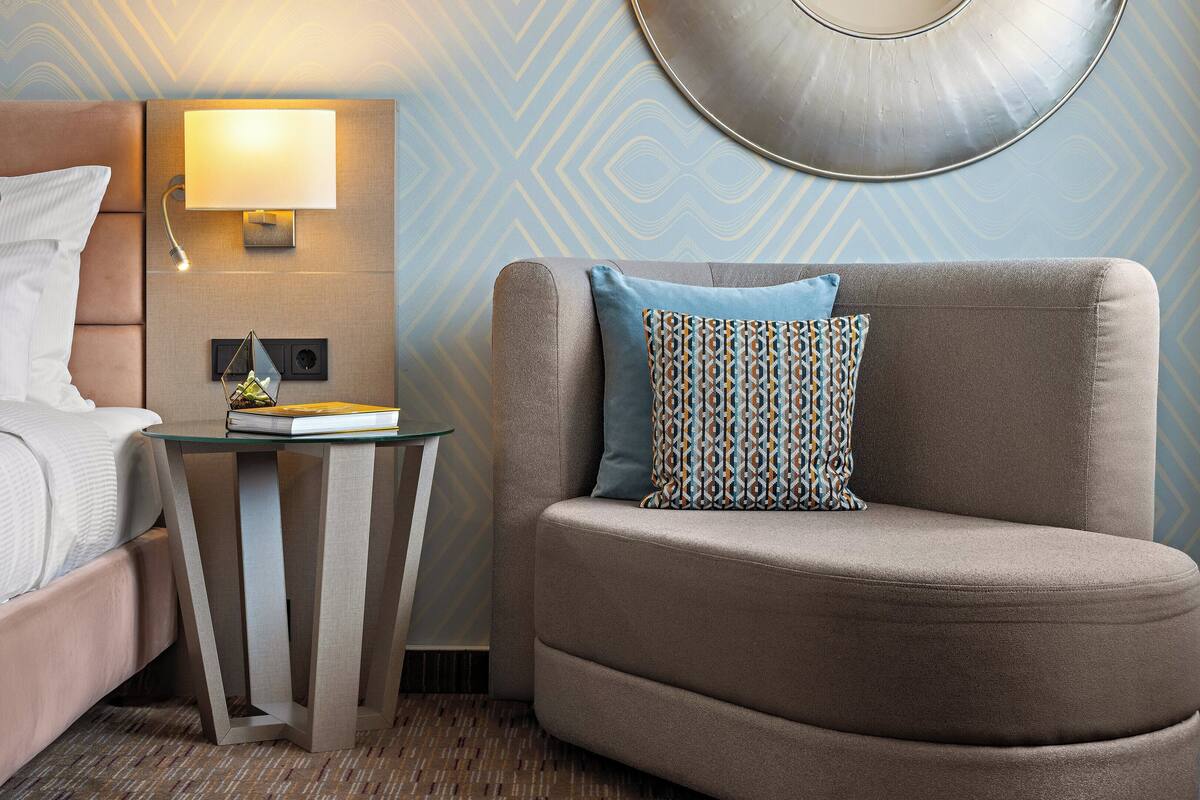 Una habitación de hotel con una cama, una mesita de noche con lámpara, una silla con un cojín estampado y una pared con diseño geométrico.