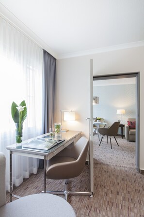 Habitación de hotel con escritorio, silla, lámpara y vista a otra habitación con cama y sillón.