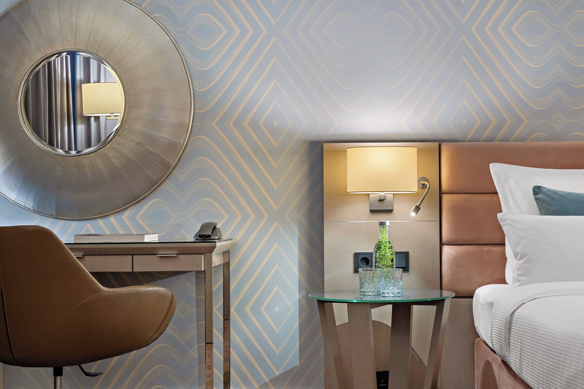Una habitación de hotel con cama, escritorio, silla, espejo redondo, lámpara de pared y una mesa de vidrio con una botella.