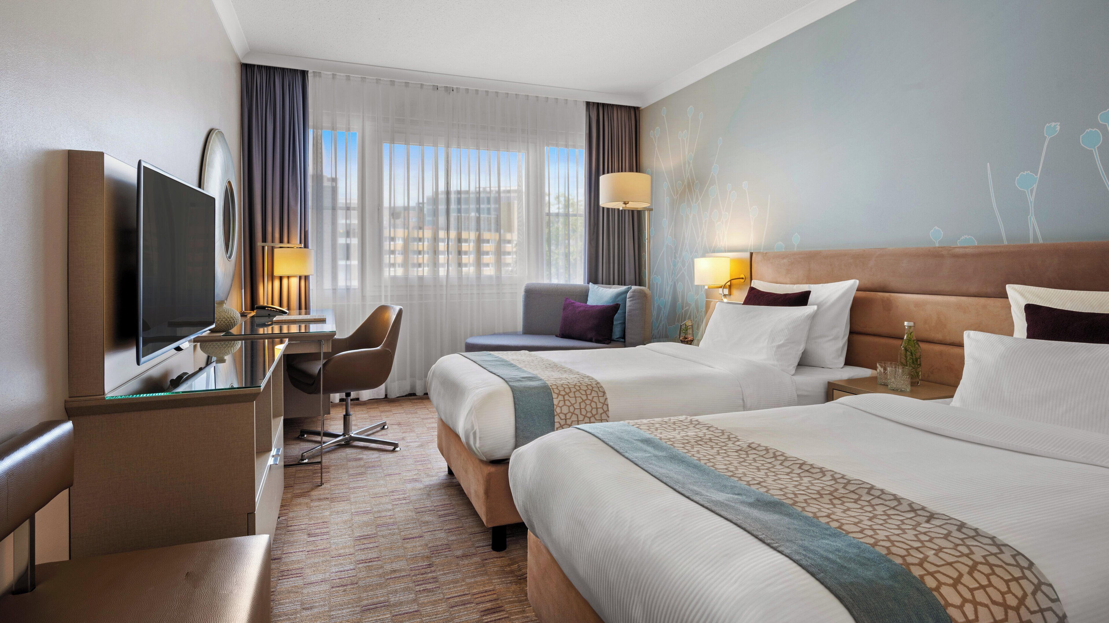 Habitación de hotel con dos camas, un escritorio con televisor, una silla y vistas a la ciudad a través de amplias ventanas.