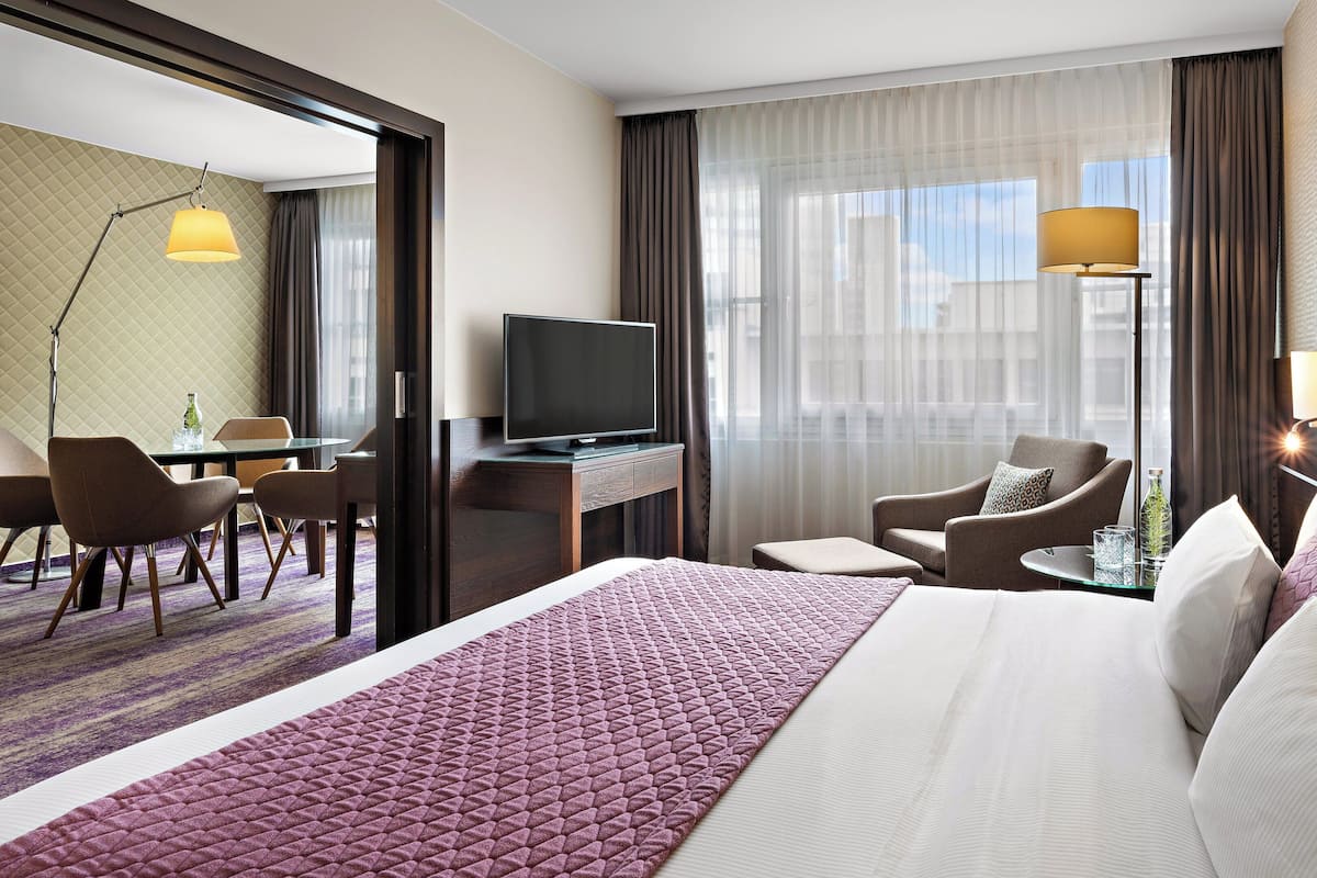 Habitación de hotel con una cama grande, un escritorio con televisor, un área de descanso con sillas y vistas a la ciudad a través de amplias ventanas.