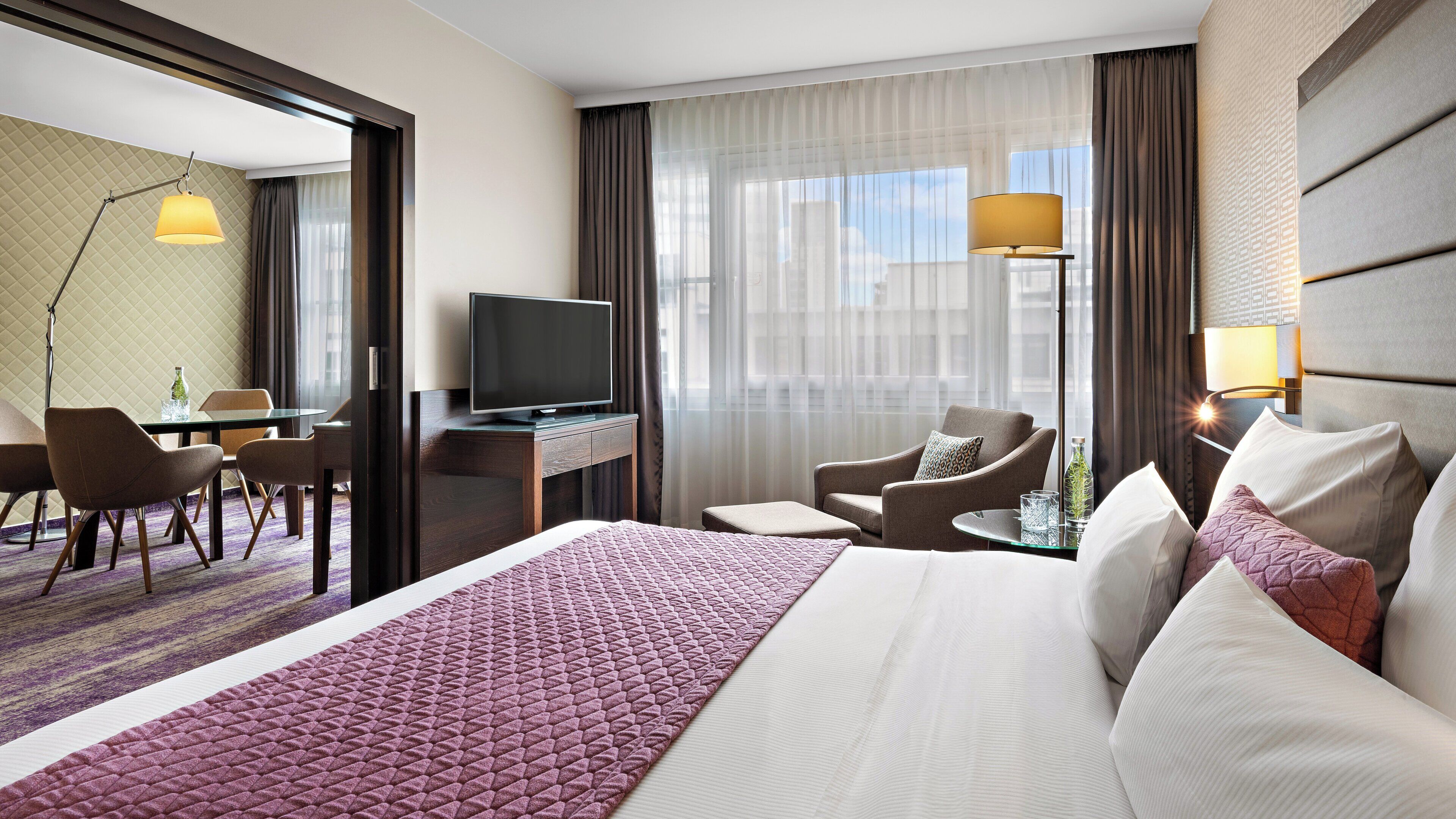 A hotel room with a large bed, a desk with a television, a seating area with chairs, and a view of the city through large windows.