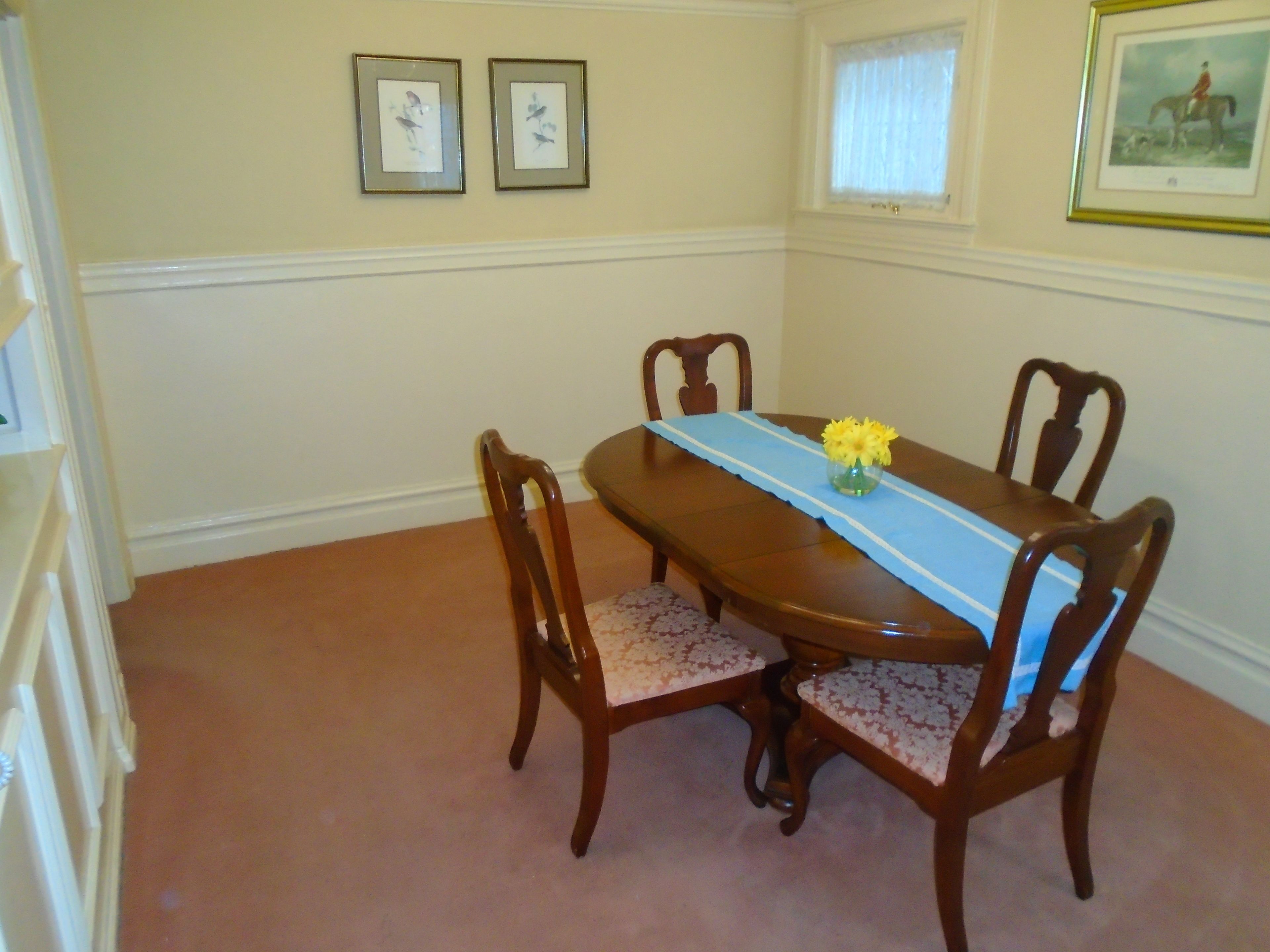 A dining room with a wooden table, four chairs, a vase with yellow flowers, and two framed pictures on the wall.