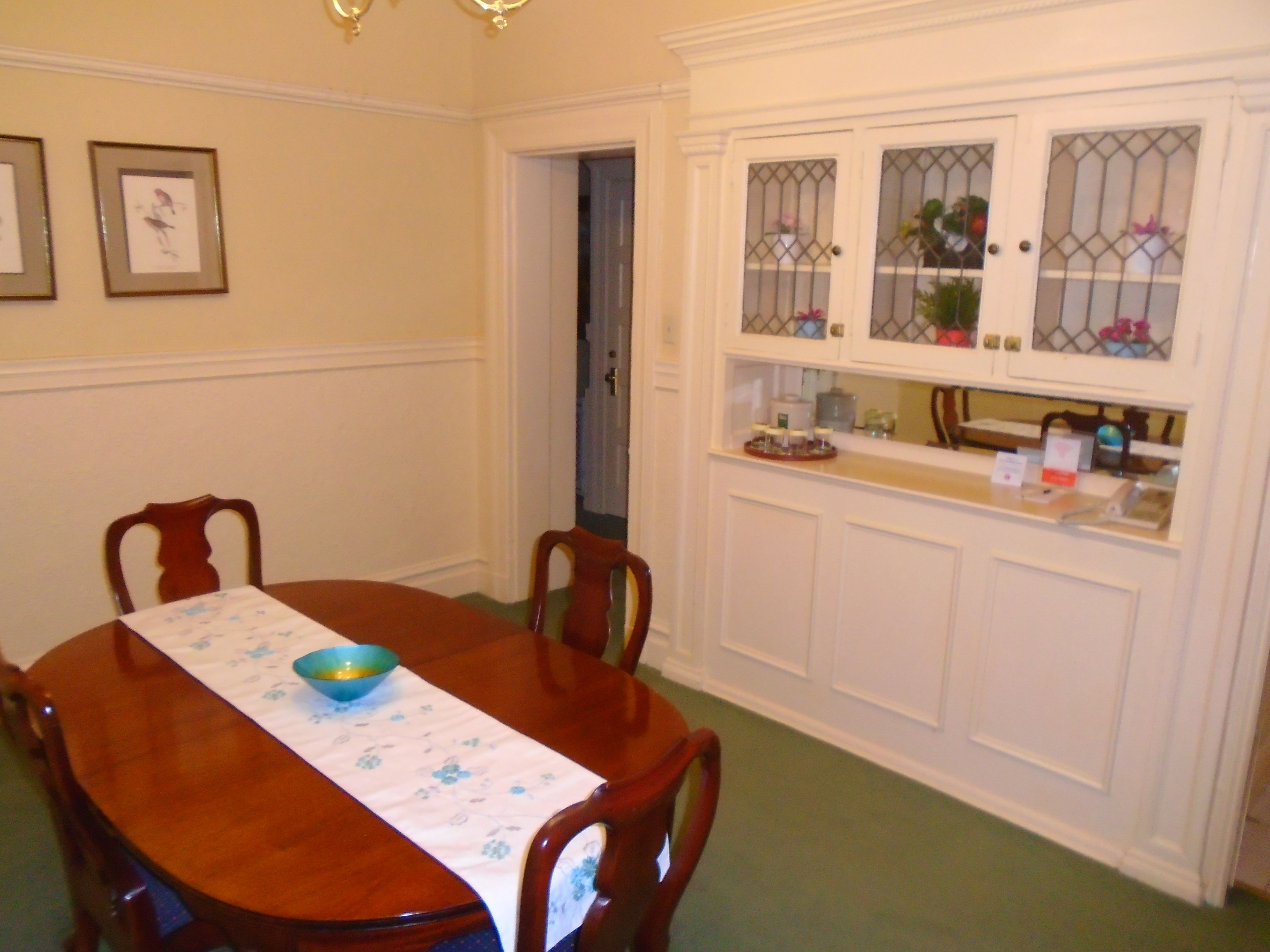 Uma sala de jantar com uma mesa de madeira, cadeiras, toalha de mesa florida e um armário branco com portas de vidro.