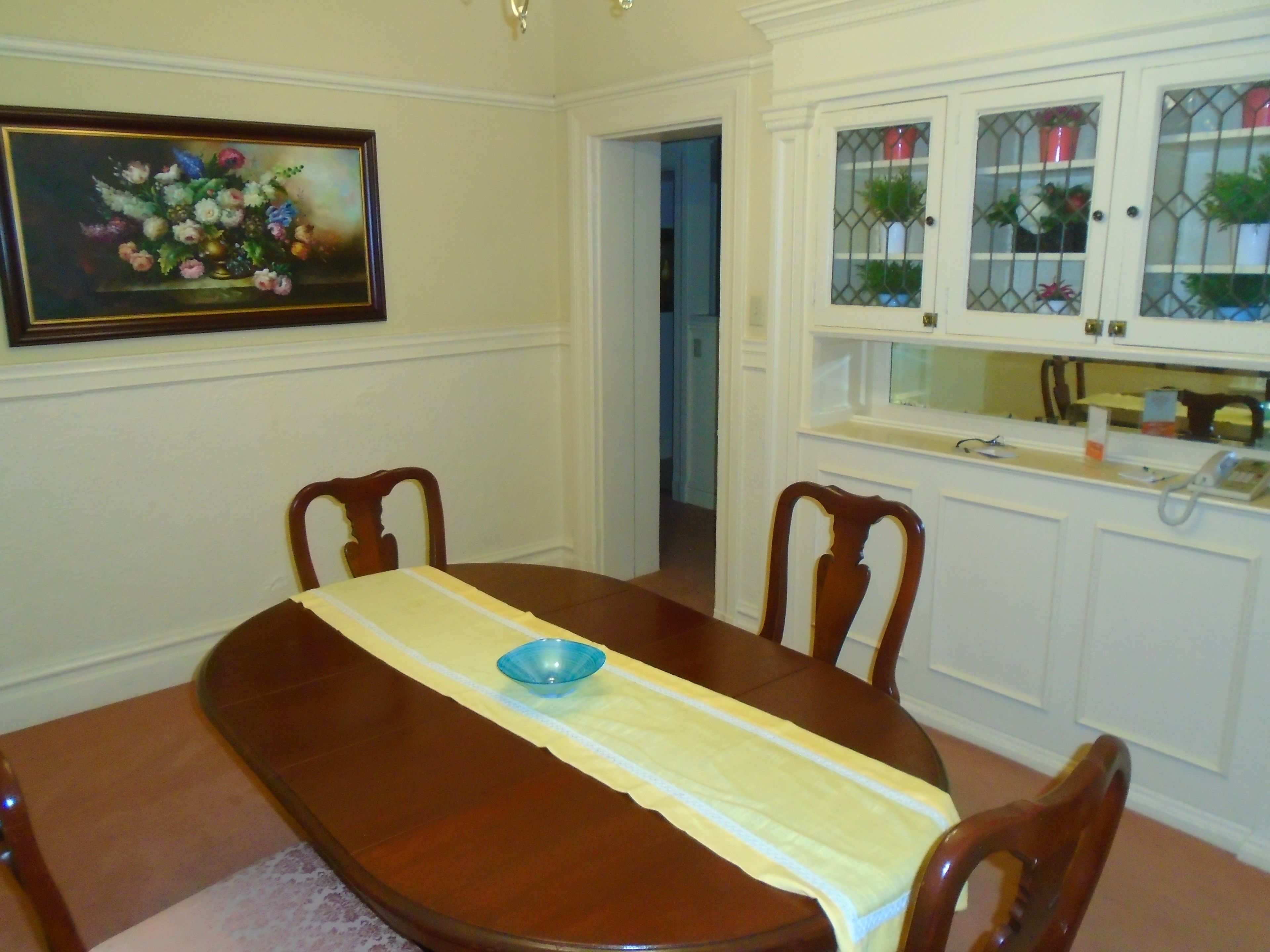 Un comedor con una mesa de madera, sillas, un cuadro y una cocina con armarios y un fregadero.