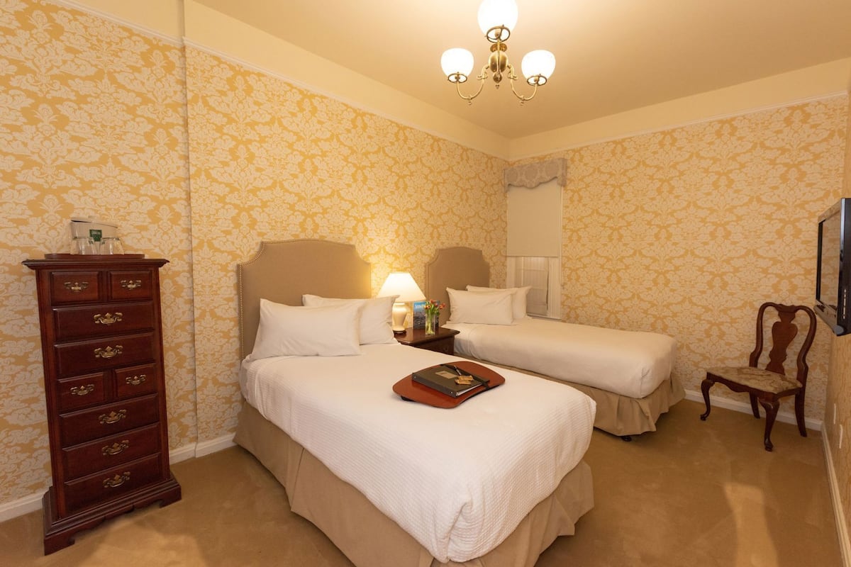 Ein Hotelzimmer mit zwei Betten, einem Schminktisch, einer Lampe, einem Sessel und einem Fernseher.