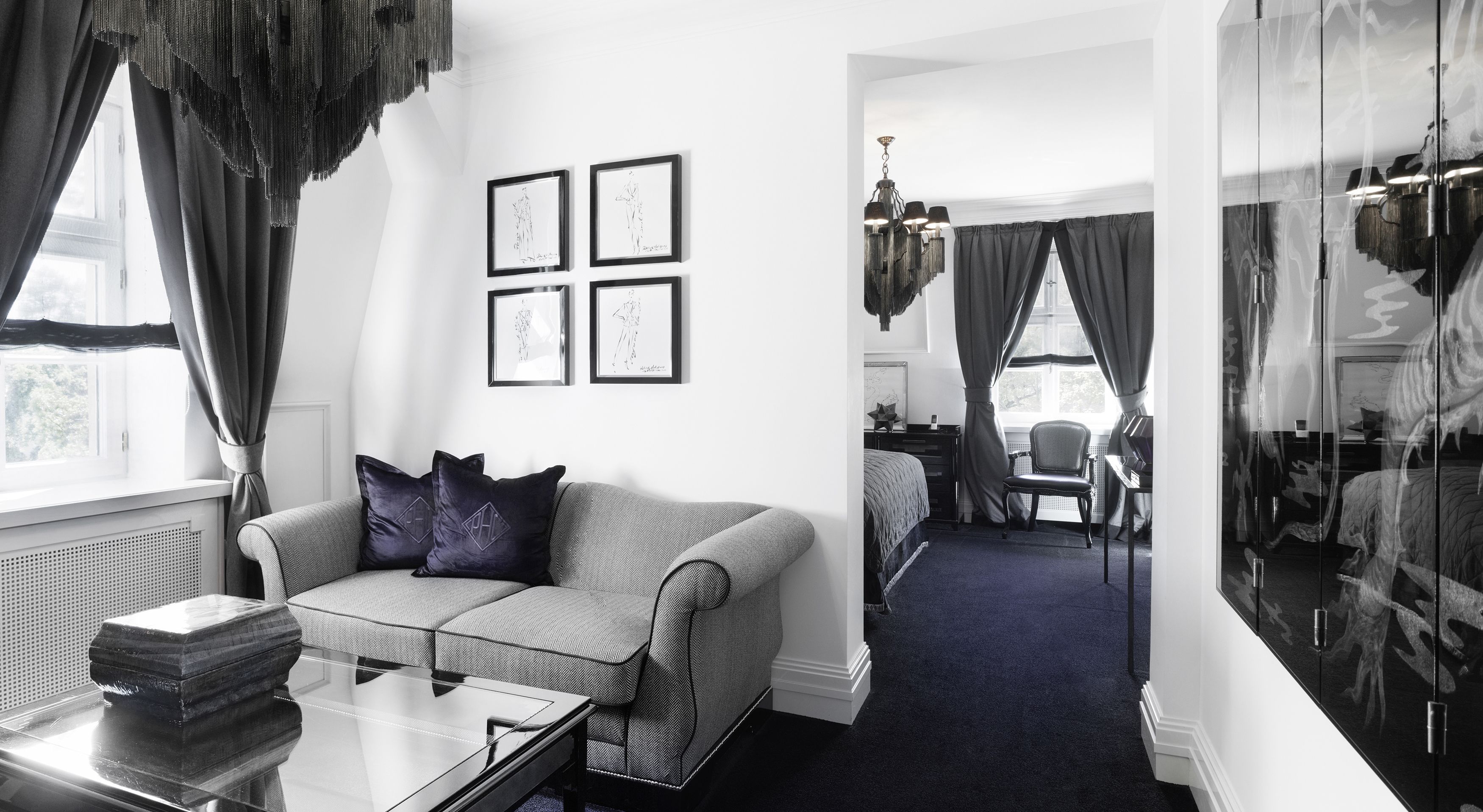 Una habitación de hotel moderna con un sofá gris, una mesa de centro de vidrio y una cama sobre una alfombra azul oscuro. Hay cuadros enmarcados en la pared y un espejo grande que refleja la habitación.