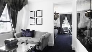 Una habitación de hotel moderna con un sofá gris, una mesa de centro de vidrio y una cama sobre una alfombra azul oscuro. Hay cuadros enmarcados en la pared y un espejo grande que refleja la habitación.