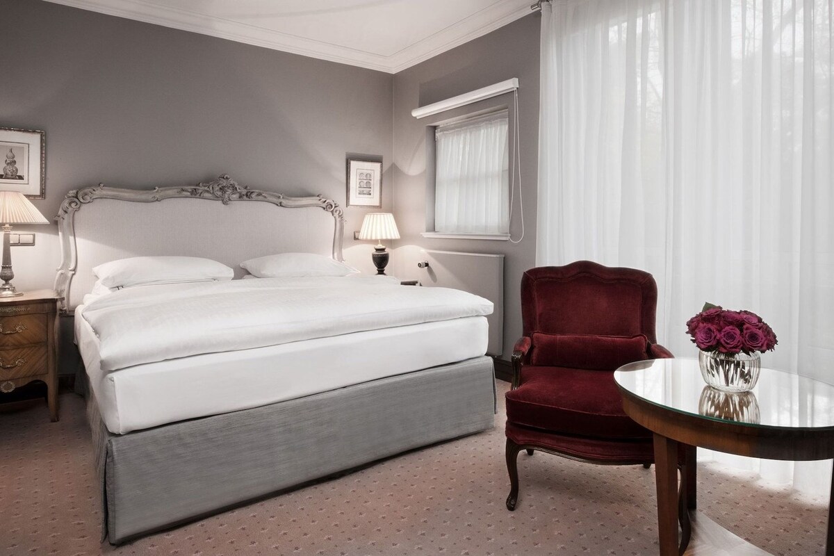 Habitación de hotel ordenada con cama, mesita de noche con lámpara, una silla roja, una mesa de cristal con un jarrón de flores y una ventana con cortinas traslúcidas.
