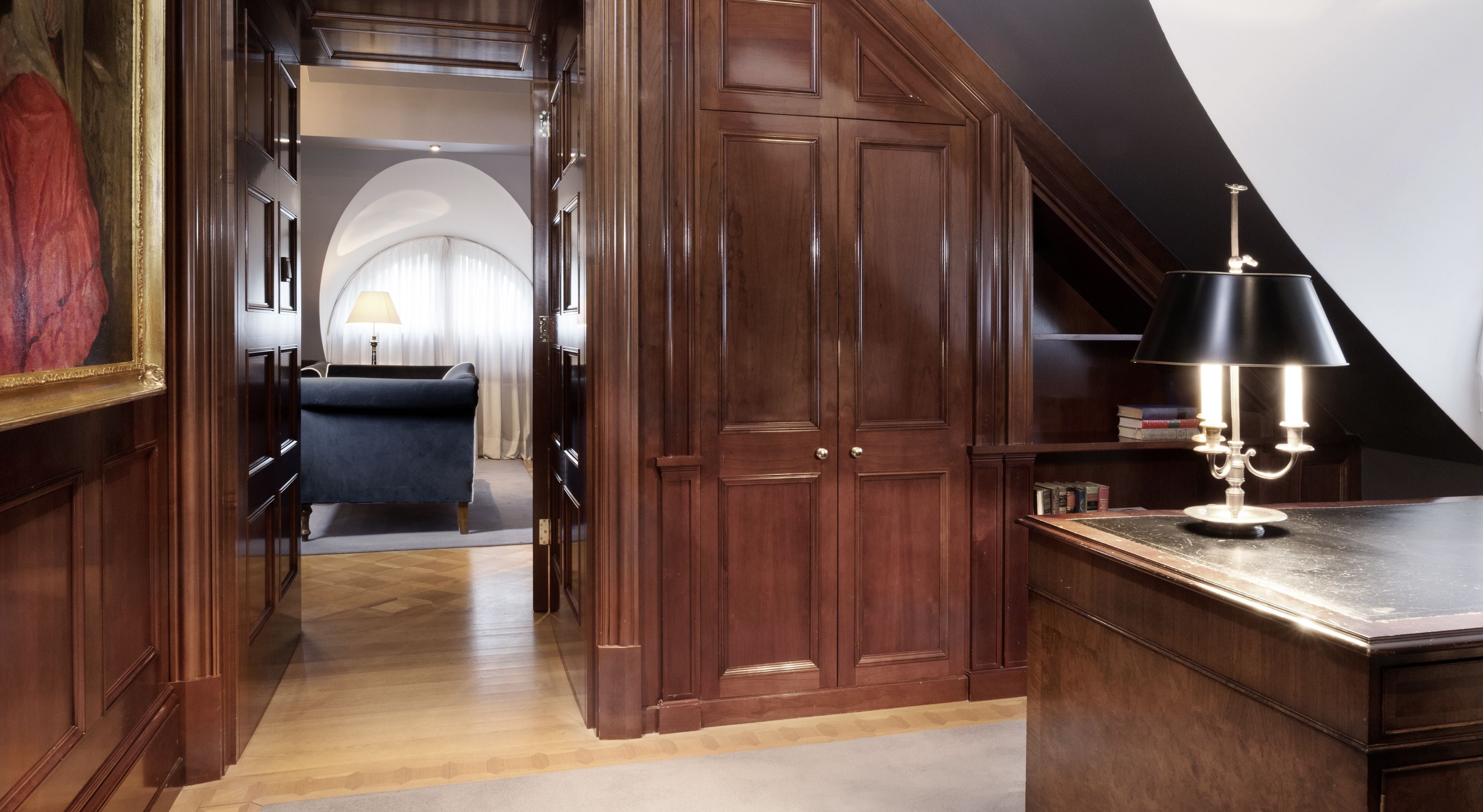 A room with wooden paneling, a desk with a lamp, a bookshelf, and a view of a living area with a couch and curtains.