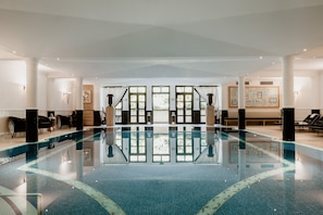A large indoor swimming pool with clear water, surrounded by white columns and seating areas.