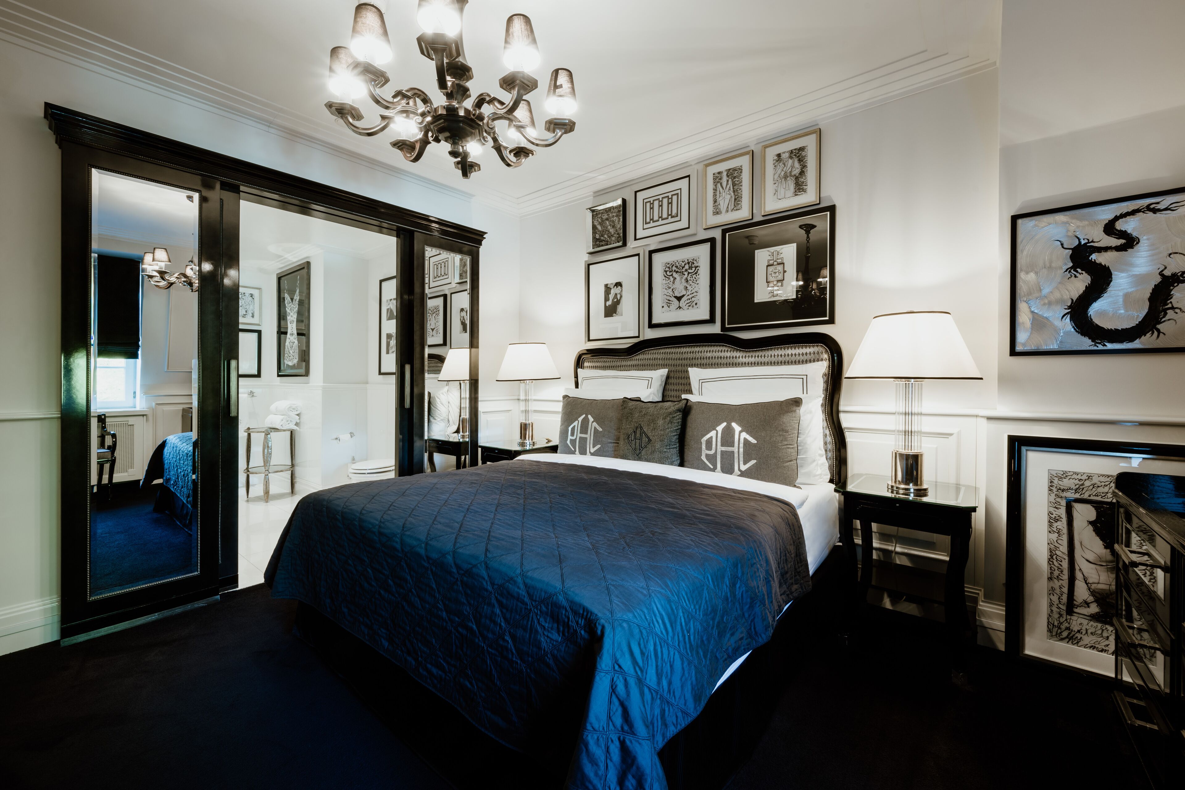 A well-lit bedroom with a large bed, a chandelier, framed artwork on the walls, and a mirror reflecting the room.