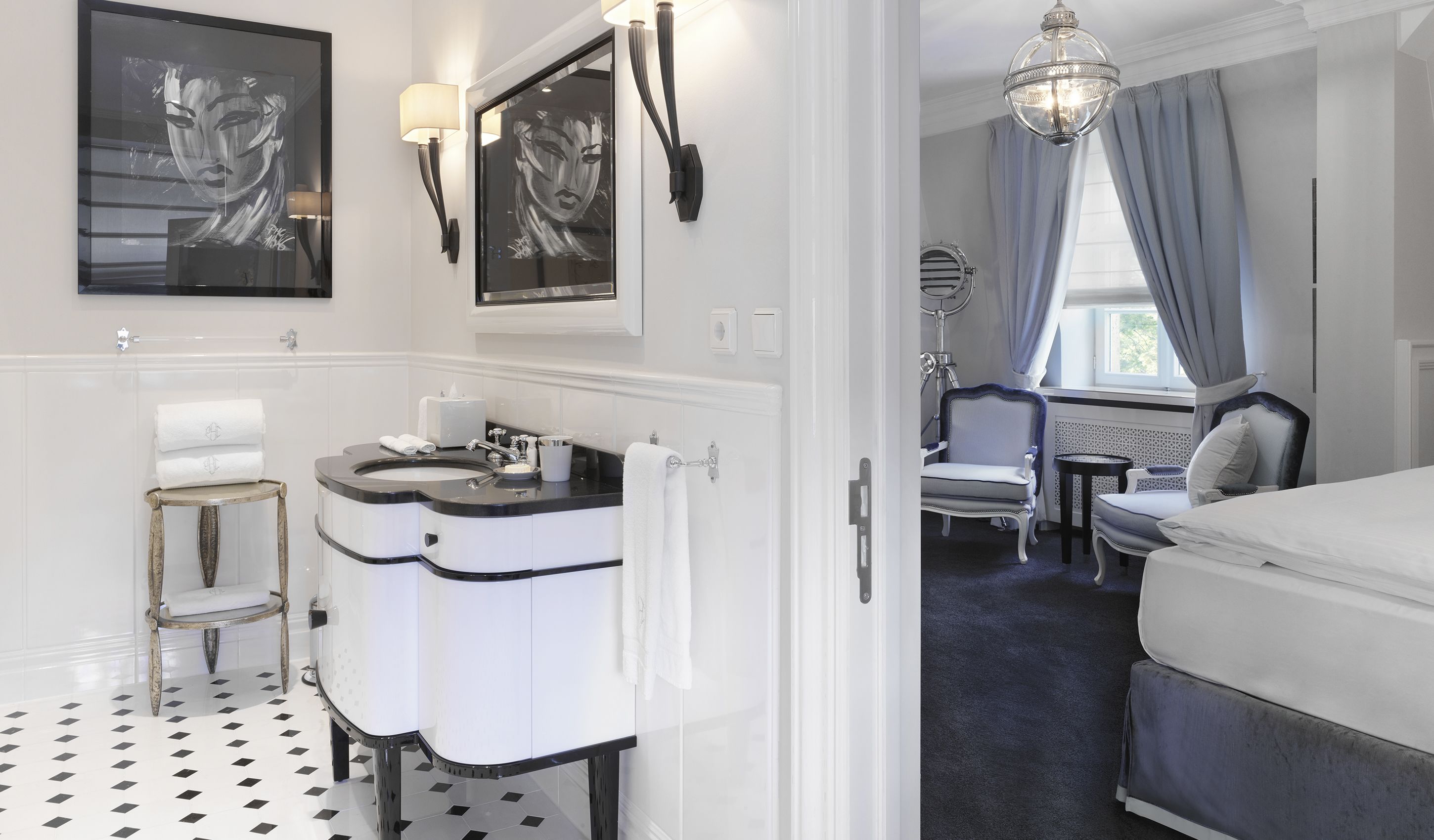Ein modernes Badezimmer mit einem Doppelwaschbecken, Waschtisch und einem Spiegel. Im angrenzenden Schlafzimmer befindet sich ein Sessel und ein kleiner Tisch mit einer Lampe.