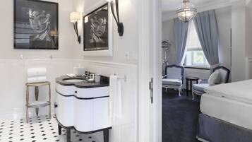 Ein modernes Badezimmer mit einem Doppelwaschbecken, Waschtisch und einem Spiegel. Im angrenzenden Schlafzimmer befindet sich ein Sessel und ein kleiner Tisch mit einer Lampe.