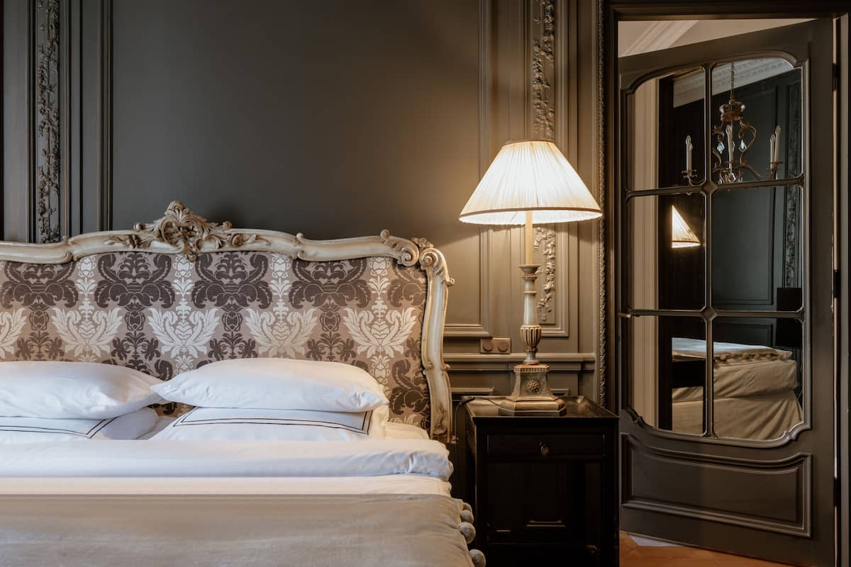 Un dormitorio con una cama grande y elaborada, una mesita de noche con lámpara y un espejo que ofrece vistas a otra habitación.