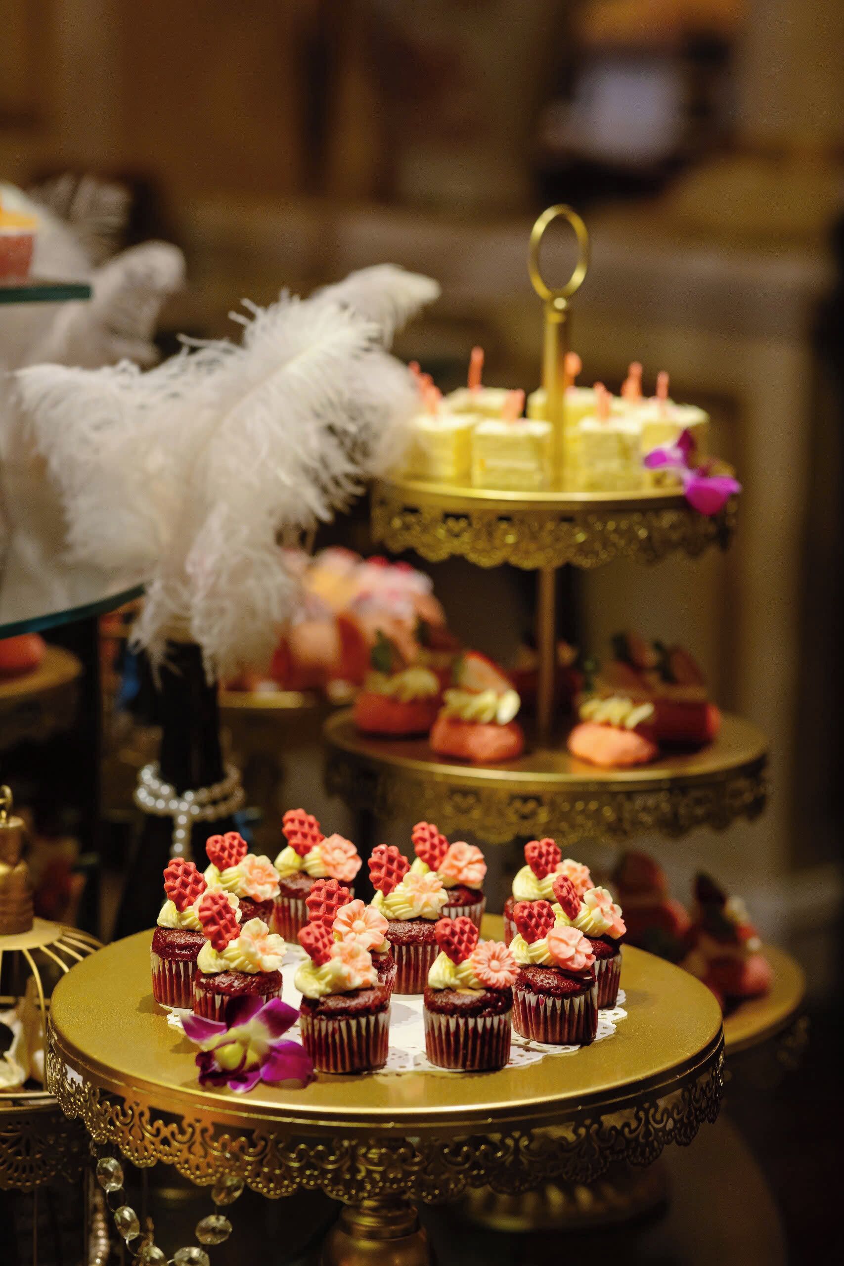 A tiered dessert stand with cupcakes and pastries, surrounded by ornate tableware and a feathered decoration.