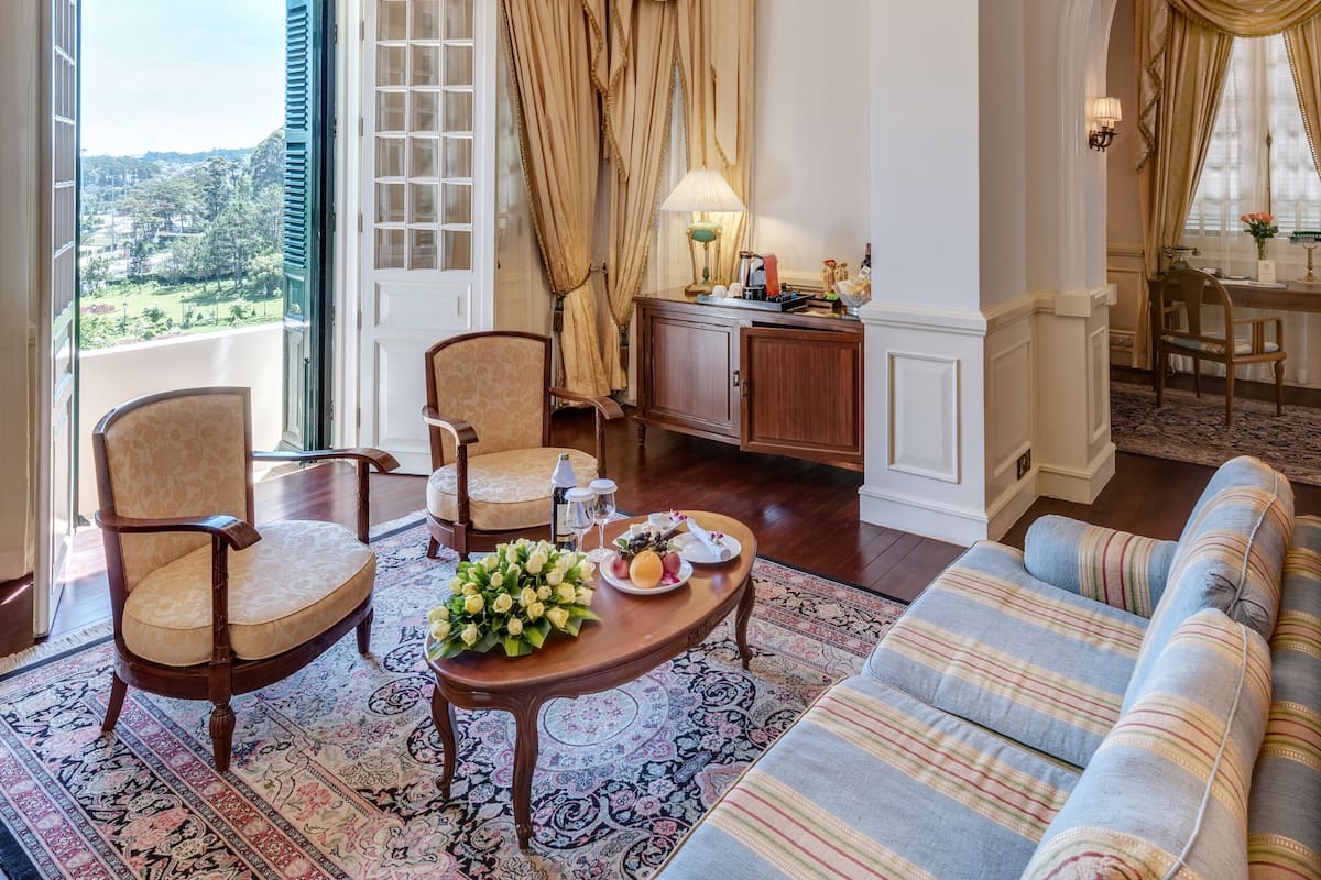 Een ruime woonkamer met een grote bank, twee fauteuils, een salontafel en een kroonluchter. Er is een eethoek met een tafel en stoelen, en een uitzicht op de omgeving door een groot raam.