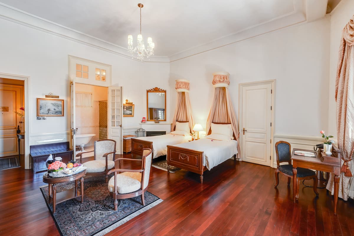Een hotelkamer met twee bedden, een zithoek met een fauteuil en een tafeltje, een bureau met een computer en een kroonluchter.