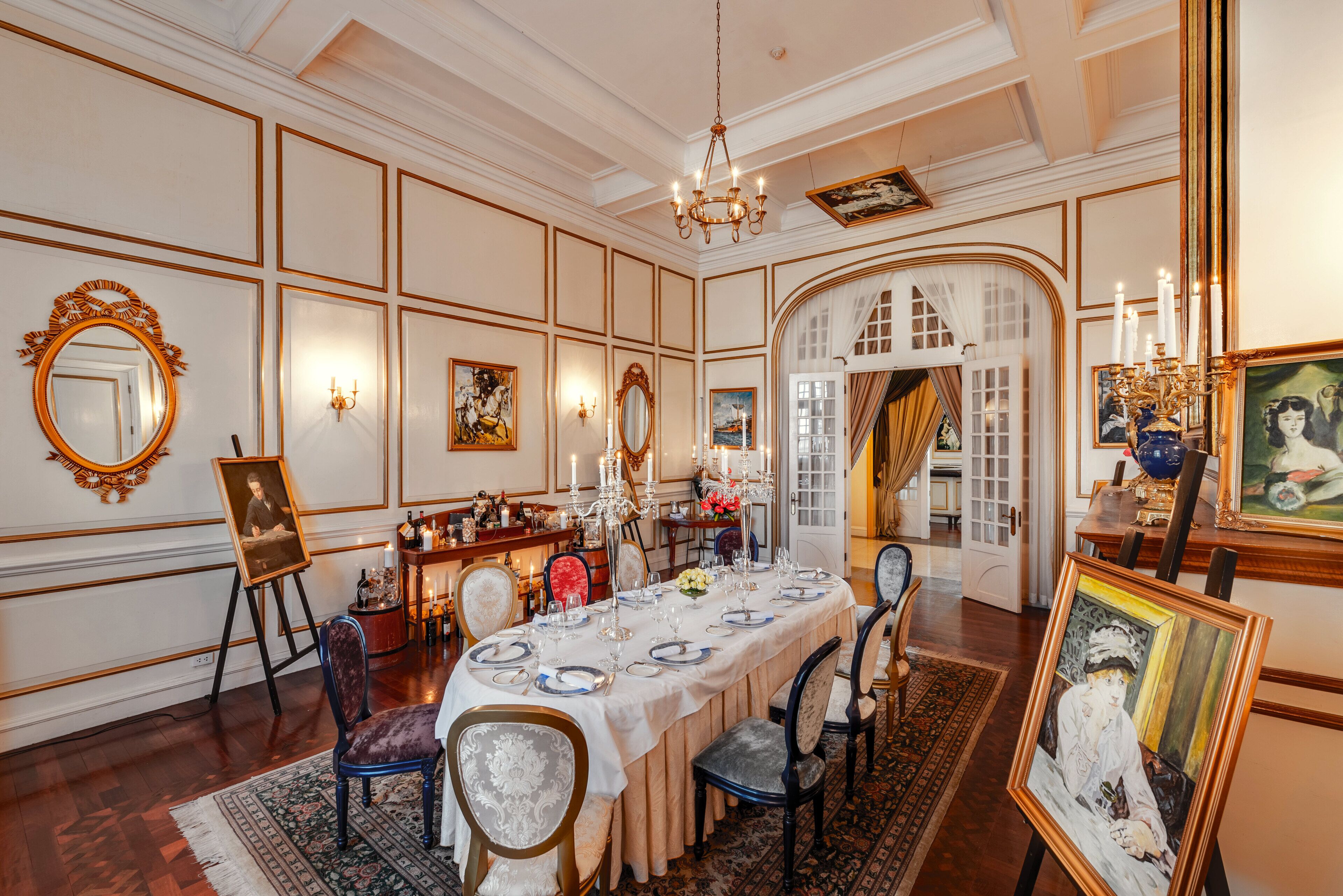 A formal dining room with a long table set for six, surrounded by ornate chairs, a chandelier, and framed artwork on the walls.