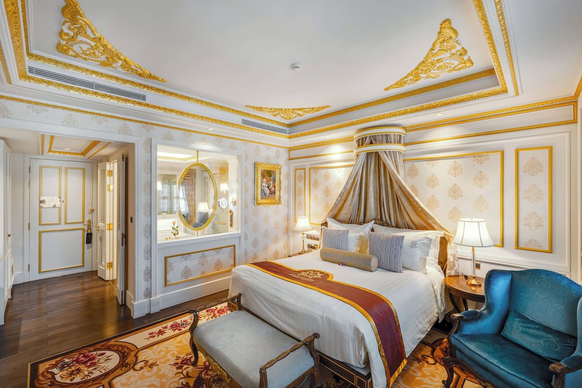 Een luxueuze slaapkamer met een groot bed, een hemelbed, een blauwe fauteuil en gedetailleerde gouden versieringen aan de muren en het plafond.