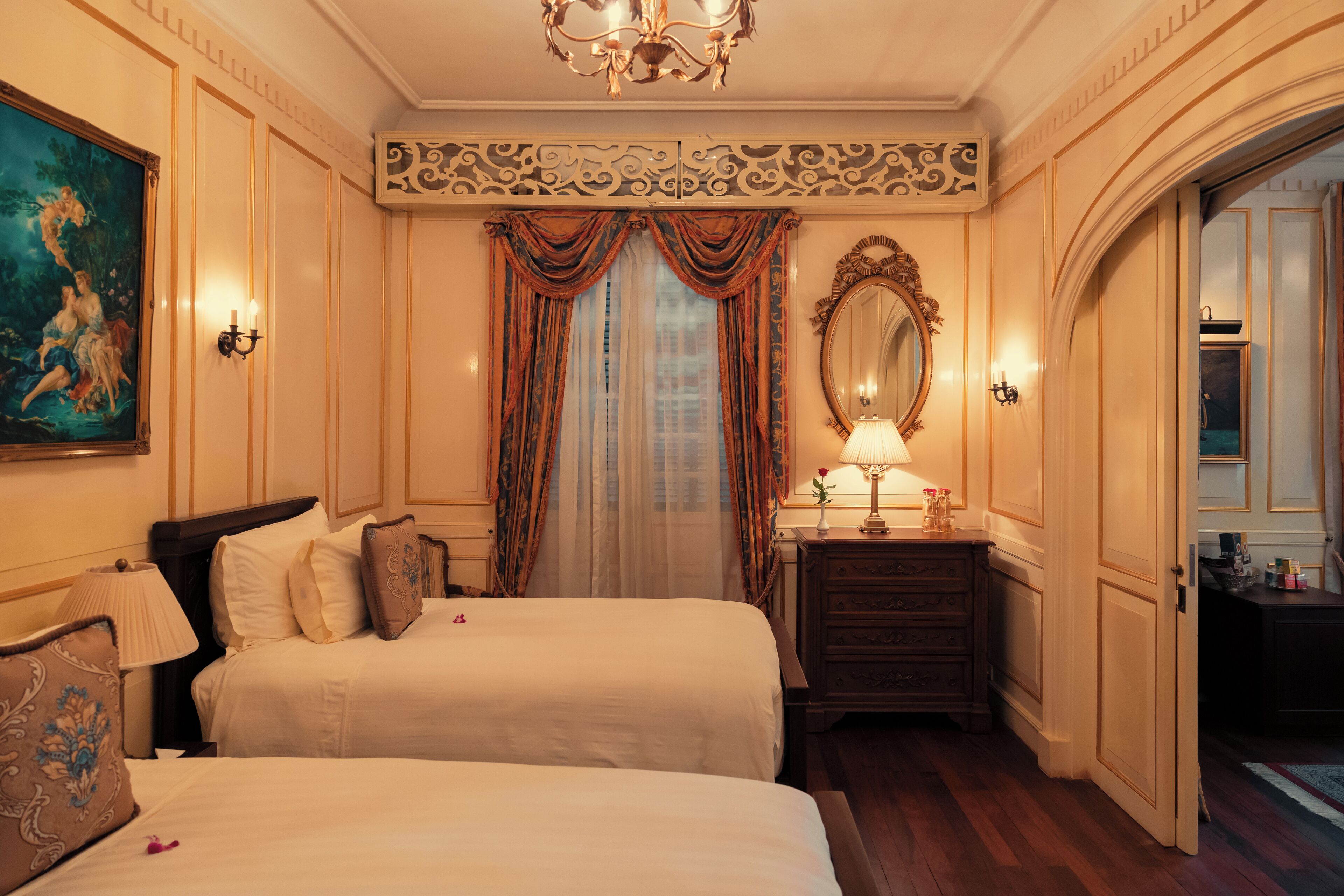 A hotel room with two beds, a large mirror, a chandelier, and a painting on the wall.