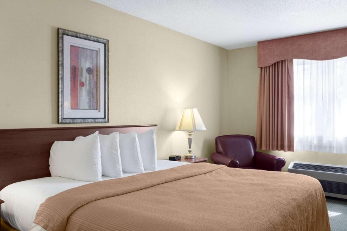 A hotel room with a bed, a chair, a lamp, a picture on the wall, and a window with curtains.