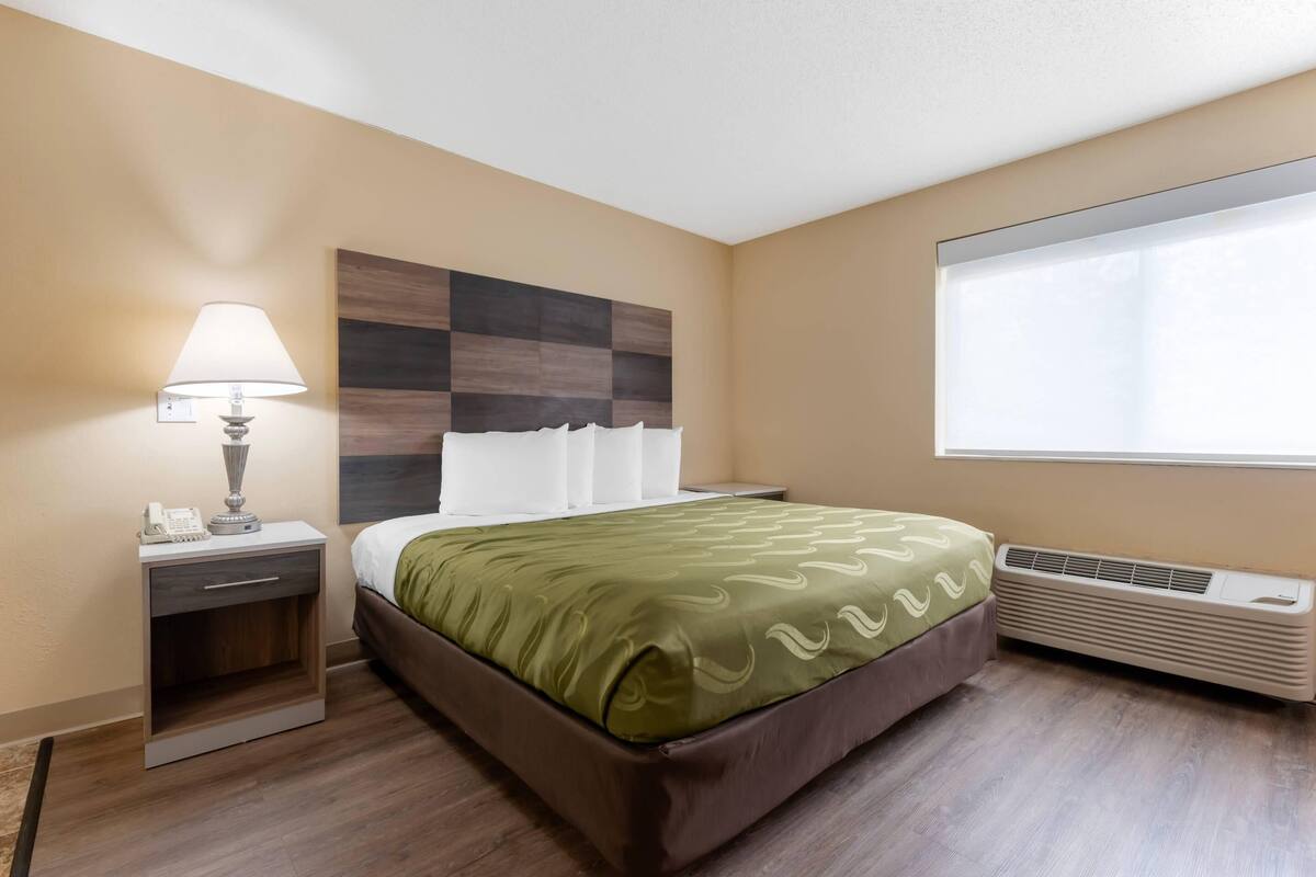 A hotel room with a bed, bedside table with a lamp, a wall-mounted air conditioner, and a window with blinds.