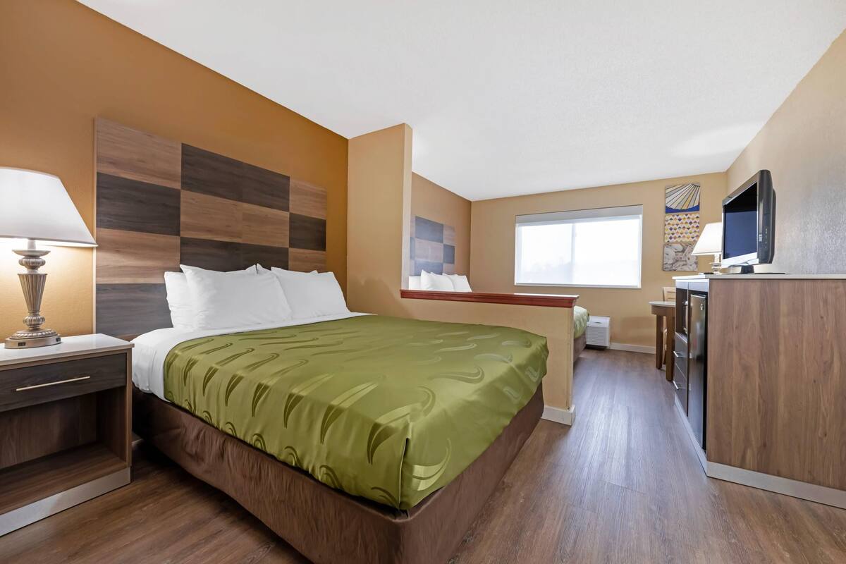 A hotel room with a bed, bedside table, lamp, and a kitchenette area.