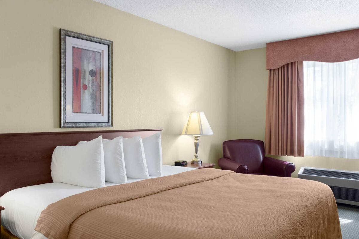 A hotel room with a bed, a lamp, a chair, a picture on the wall, and an air conditioning unit.