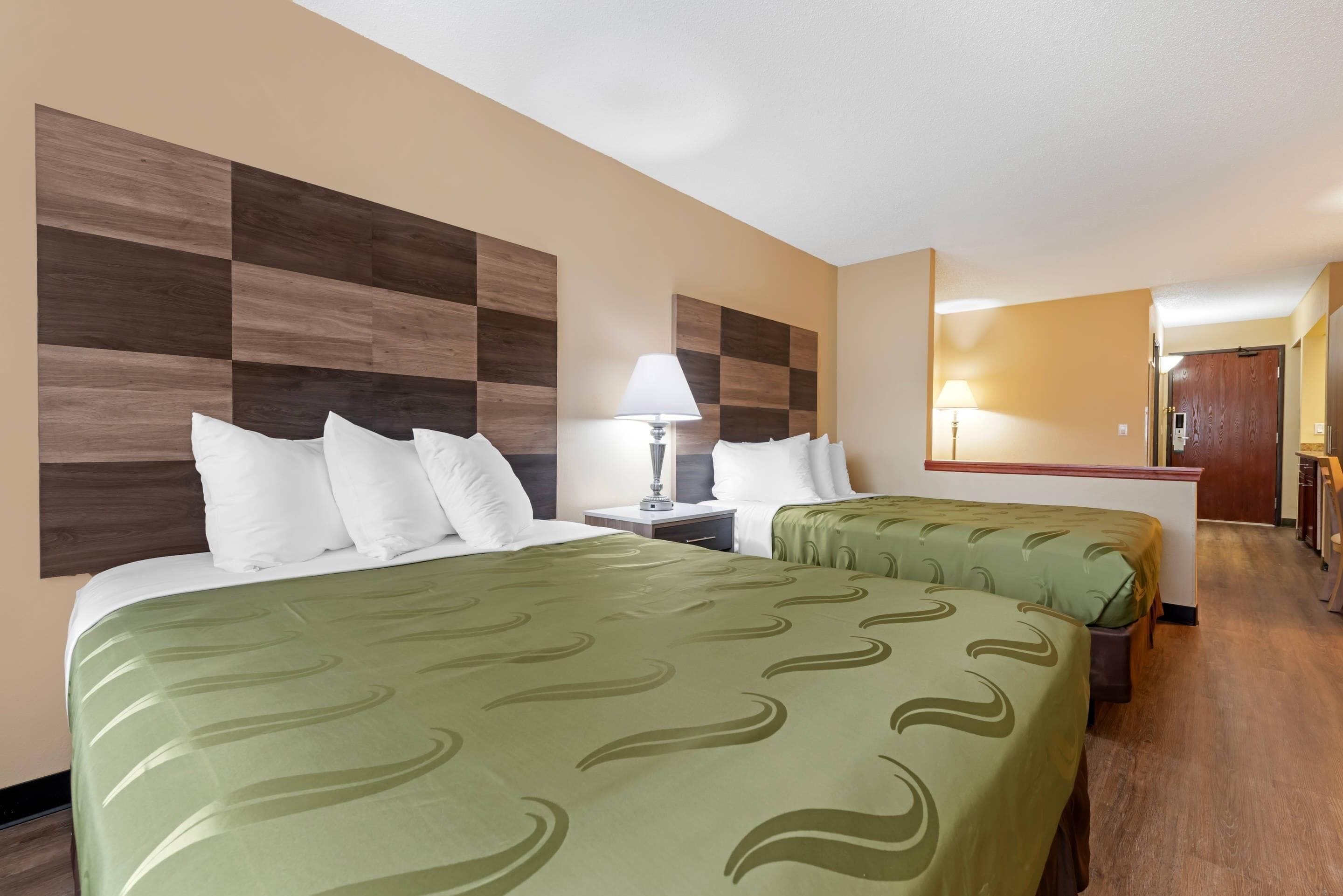 A hotel room with two beds, a nightstand with a lamp, a wooden headboard, and a view of the hallway.