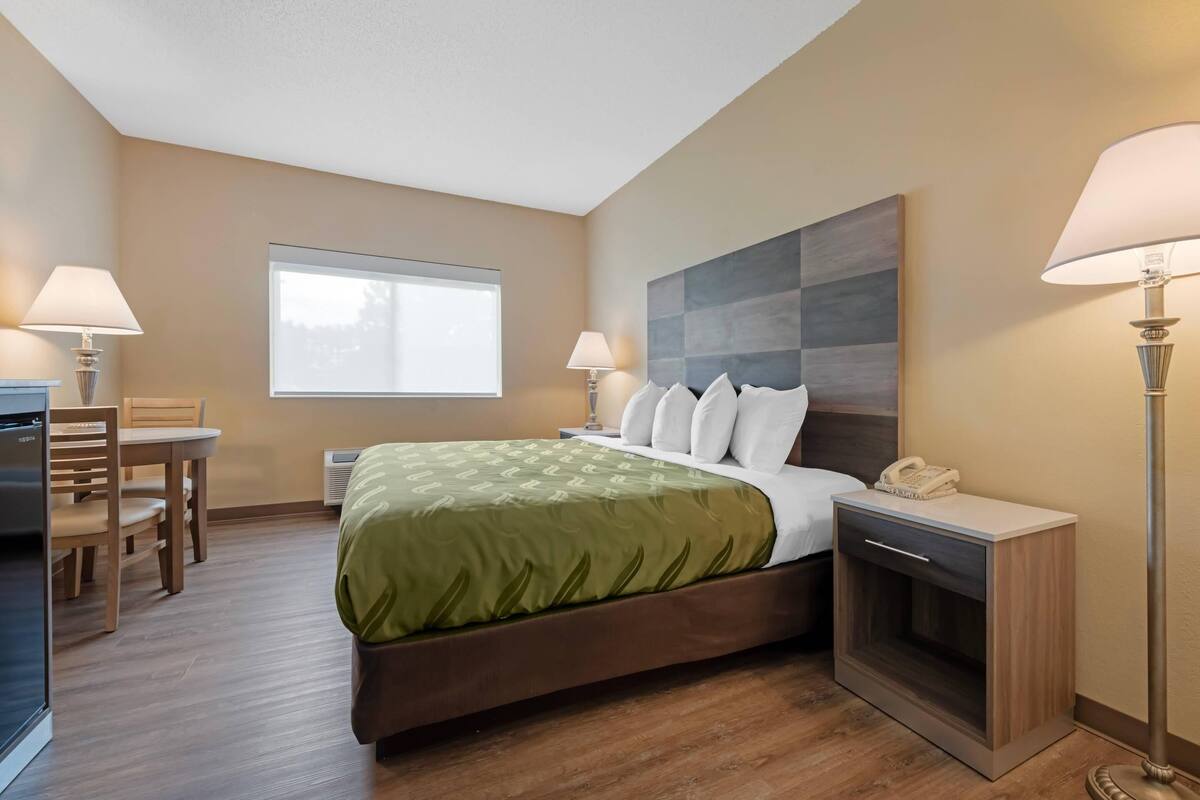 A hotel room with a bed, bedside table, lamp, desk, chair, and a window.