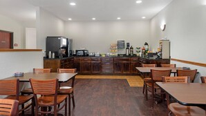 Un comedor de hotel con mesas y sillas de madera, una zona de cocina con electrodomésticos y una pared blanca con una lámpara.